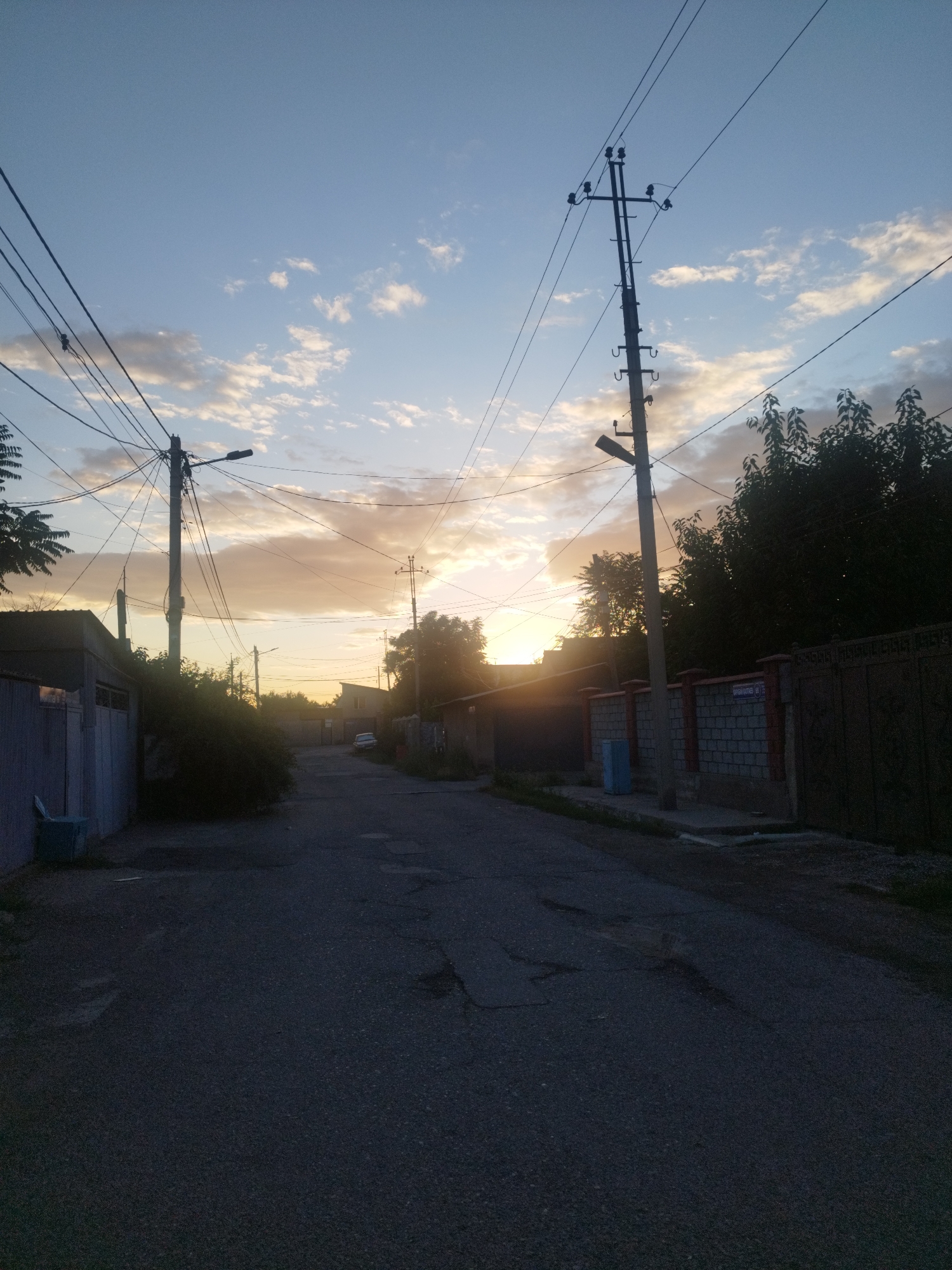 The image captures a street scene in Shymkent, Kazakhstan, during twilight. The sky is a pale blue with wispy, yellowish clouds, indicating the sun is low on the horizon. A network of electrical wires and poles dominates the upper portion of the frame, crisscrossing the sky. On either side of the street are residential buildings with fences. To the left, a blue fence and a portion of a building are visible. To the right, a brick fence with a dark metal gate stands, and further down, a car is parked on the asphalt road. The road itself is paved and shows signs of wear and tear. Trees and bushes are present on both sides of the street, some casting shadows on the road. The overall impression is of a quiet, urban residential area at the close of day.