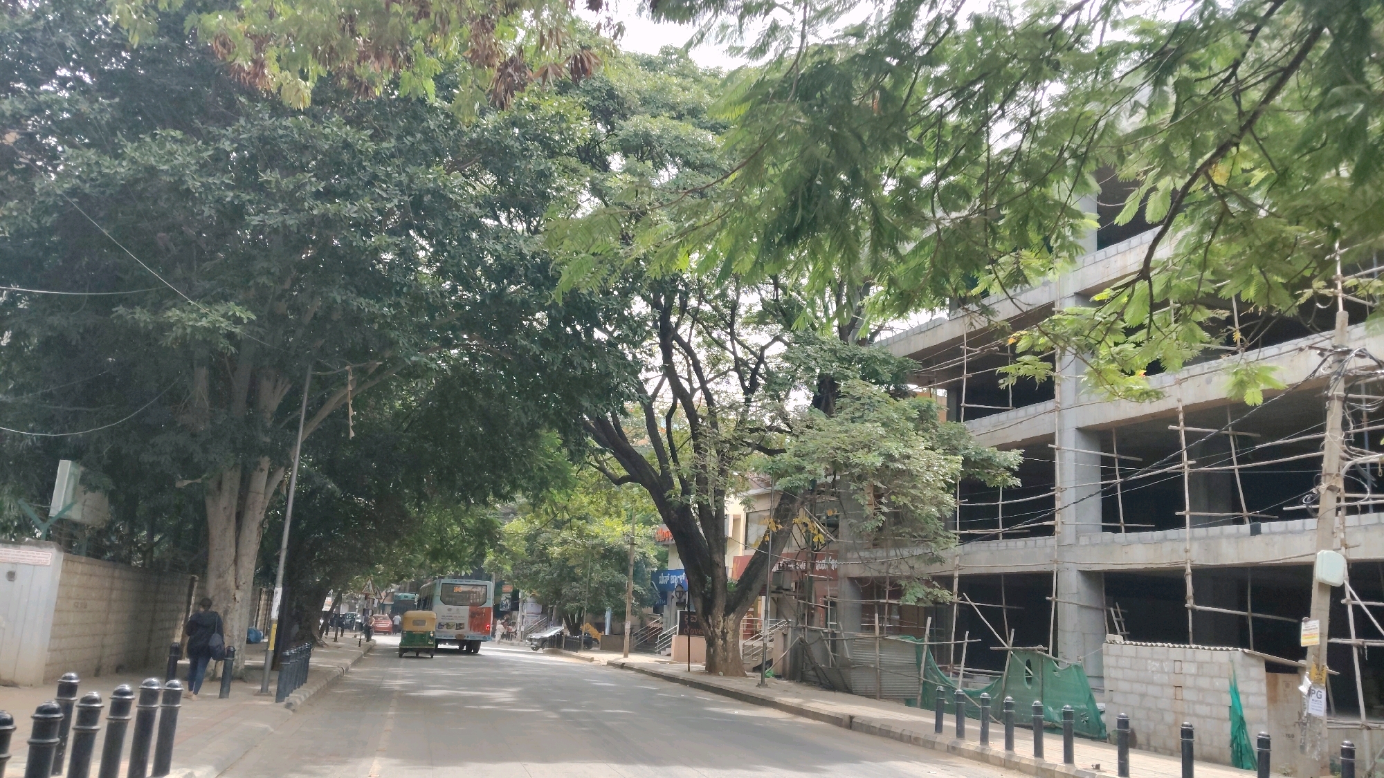 The image depicts a street scene in Bengaluru, India, likely in a commercial or residential area with ongoing construction. A multi-story building is under construction on the right side of the street, with scaffolding covering its exterior. The building appears to be made of concrete and brick. On the left side of the street, a row of trees lines the sidewalk, providing shade. A person is walking on the sidewalk. A bus and an auto-rickshaw are driving on the street, suggesting it is a public road. The sky is overcast, indicating a cloudy day. The overall atmosphere is one of urban development and daily life in a busy city.