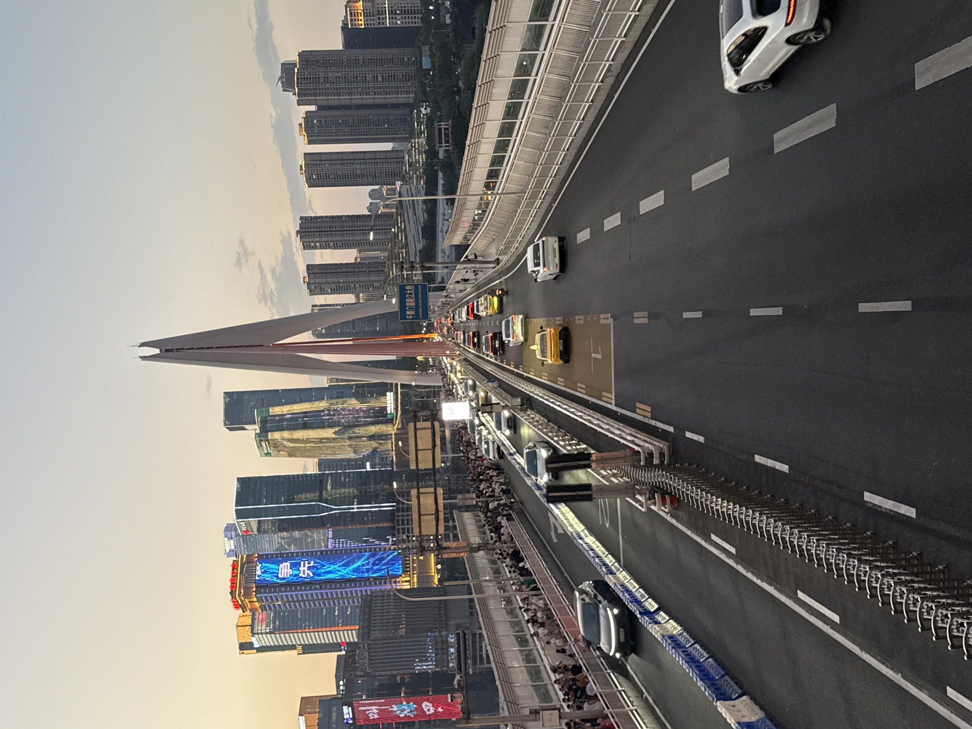 This is an eye-level, full shot of the Hongya Cave Bridge in Chongqing, Yuzhong District, China, during sunset. The bridge features a distinctive, Y-shaped suspension tower, with the iconic Hongya Cave buildings illuminated in the background. Traffic flows in both directions across the multi-lane bridge, with cars and a motorcycle visible on the roadway. Pedestrians are walking along the elevated pedestrian walkways on either side of the bridge, some with their backs to the camera, others facing the skyline. The sky is a pale orange and yellow, indicating the sun is low on the horizon. The scene captures the blend of urban infrastructure and natural light in a bustling city environment.