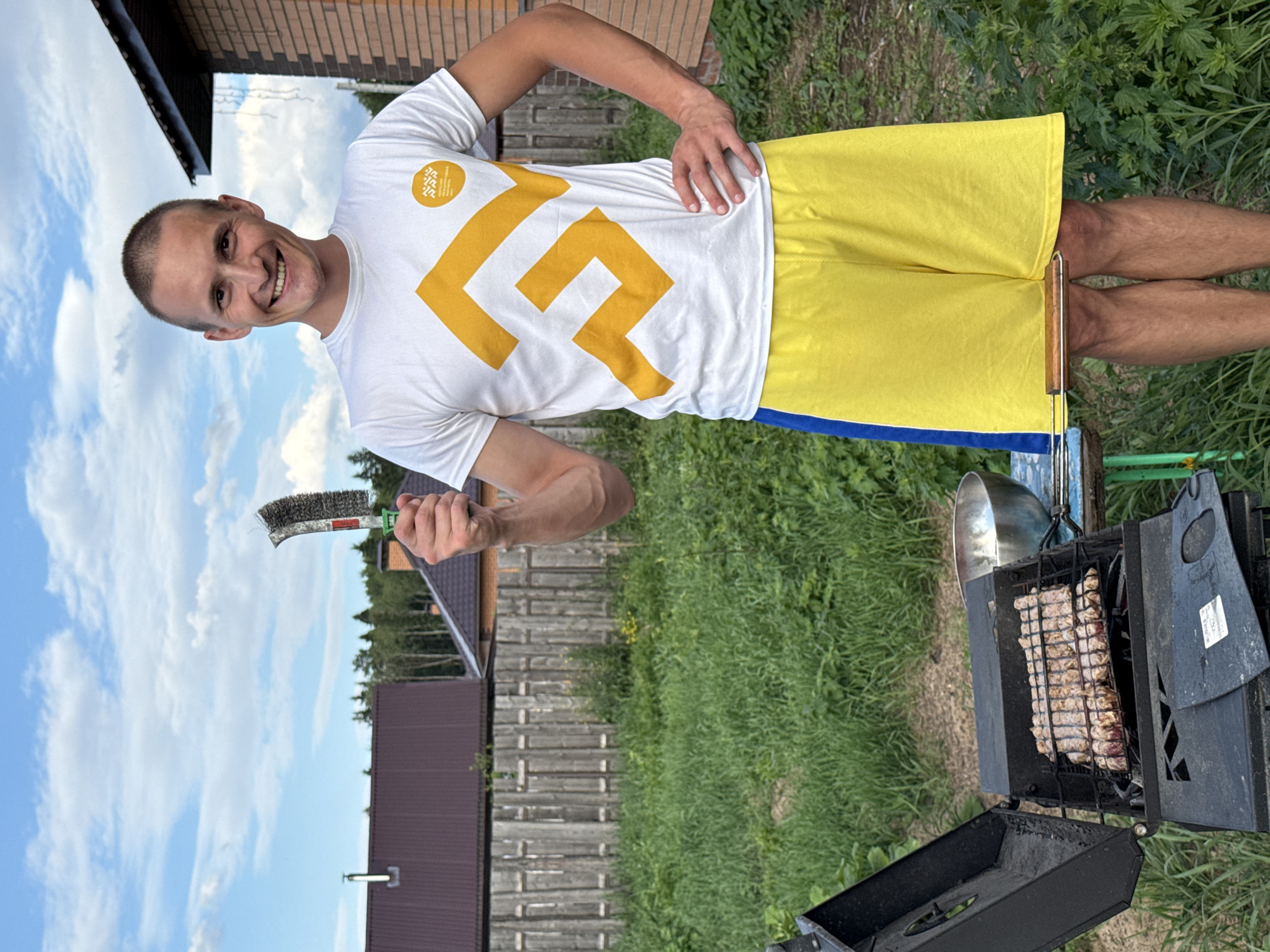 A man with short, shaved brown hair and a clean-shaven face is smiling at the camera. He is wearing a white t-shirt with a large yellow geometric design and yellow shorts with a blue stripe at the bottom. He is holding a grill brush in his right hand and his left hand is on his hip.
The man is standing outdoors in what appears to be a backyard or a park. Behind him, a grill with meat cooking on it is visible. A metal bowl hangs from the side of the grill. The setting includes a wooden fence, grass, trees, and a brick building in the background. The sky is blue with white, fluffy clouds, suggesting it is daytime and the weather is fair. The overall mood is cheerful and relaxed, indicating a leisure activity like a barbecue.
The image appears to be taken in Chaikovsky, Russia, based on the provided location context. No visible text is present on the man's clothing, the grill, or in the background. The image is oriented vertically, with the man's head on the left side and his legs on the right.