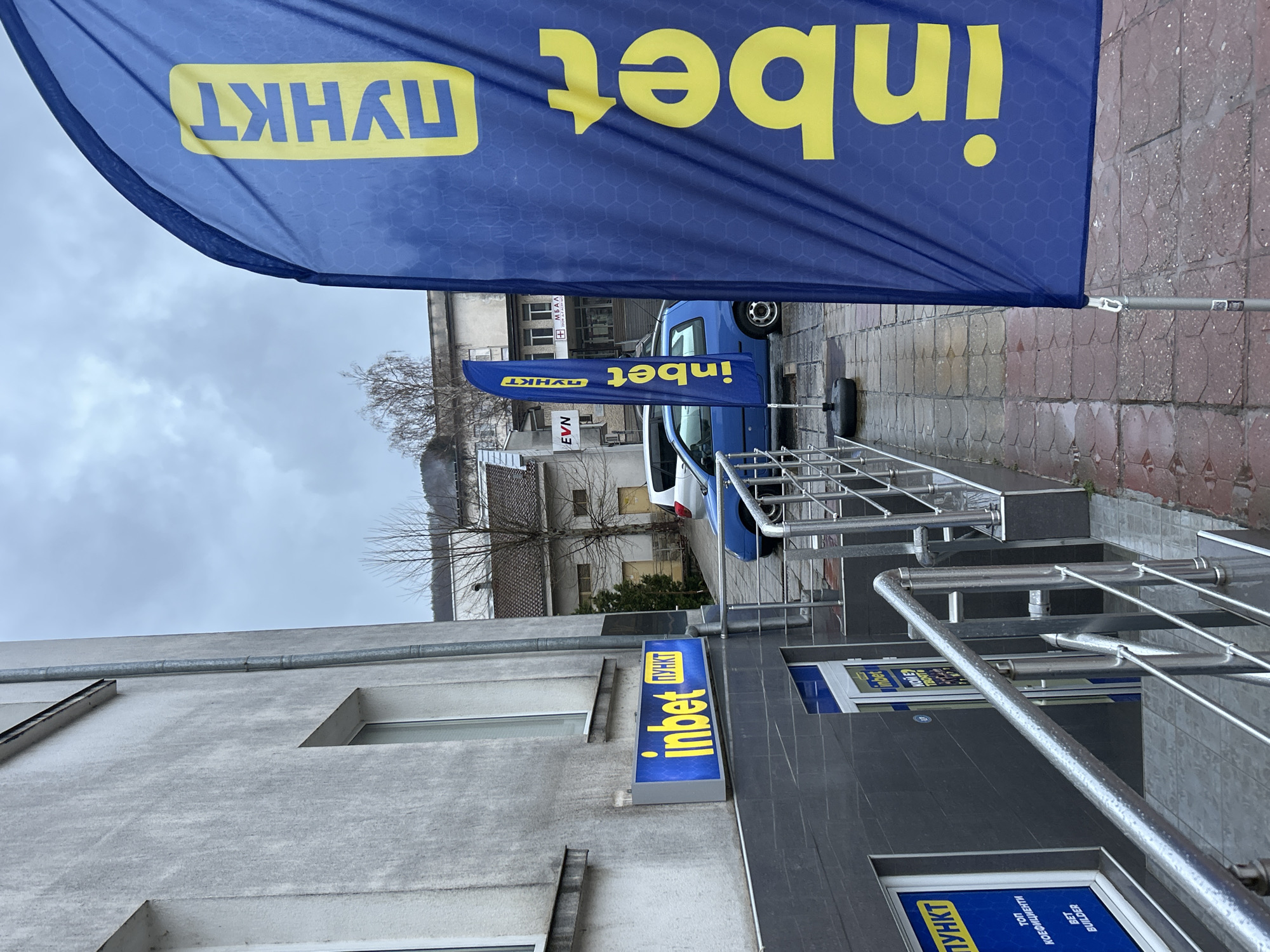 The image depicts the exterior of a "inbet" betting shop in Zlatograd, Bulgaria. Signage for "inbet" is prominently displayed, including banners and storefront signs in blue and yellow. The signage also includes the word "ПУНКТ." The image captures architectural details of the building, as well as elements of the urban environment like a vehicle, handrails, and a nearby EVN sign. The sky is overcast, and the overall scene presents an ordinary street-level view.