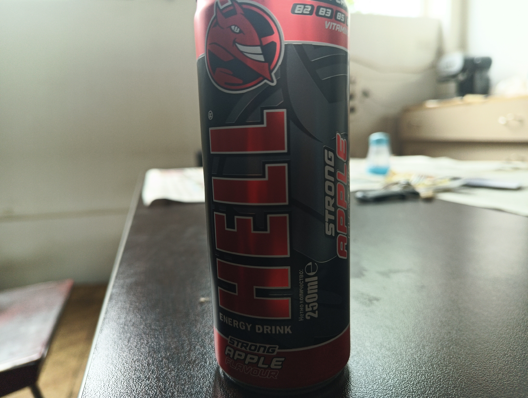 The image shows a can of "Hell" energy drink on a table. The can is red, black, and grey, with the word "HELL" in large, red letters. The flavor is "Strong Apple." The can also features a devil logo. The background is blurred, but it appears to be an indoor setting with a wall and furniture.