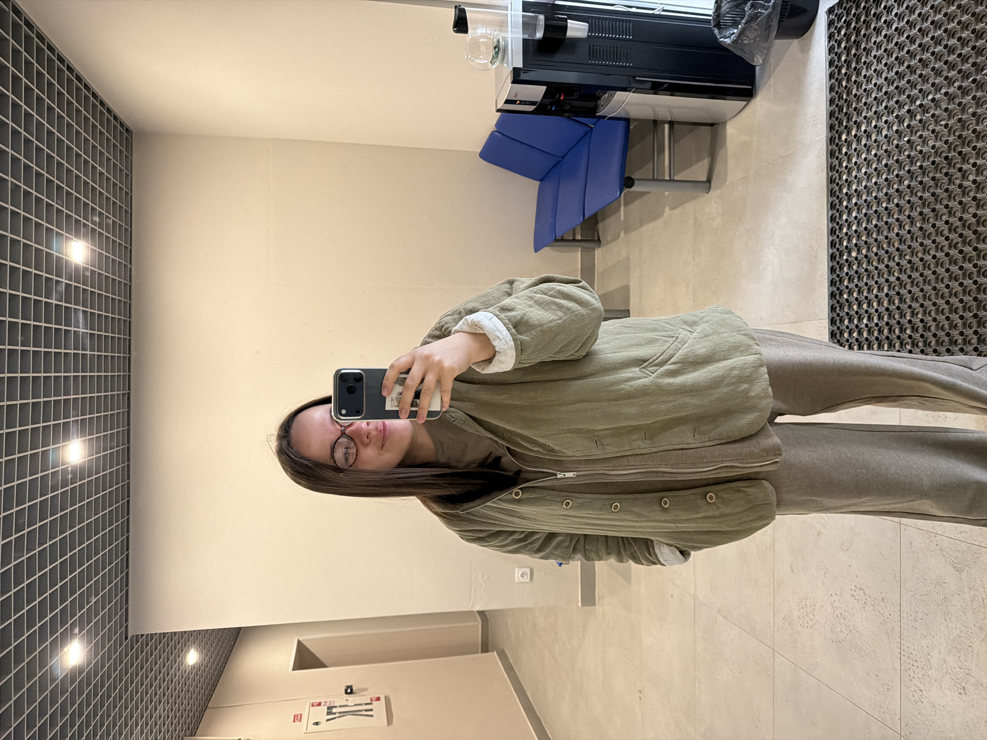 A young woman with long brown hair and glasses is taking a selfie in an interior space with light beige walls and tiled floors. She is wearing a green buttoned jacket over a beige hoodie and matching sweatpants. In the background, there is a black tile wall with lights, a blue chair, a black machine on a table, and a black floor mat. A sign with pictograms is visible on the wall. The photo was likely taken in a public building or facility in the city of Chaikovsky, Russia, as indicated in the prompt.