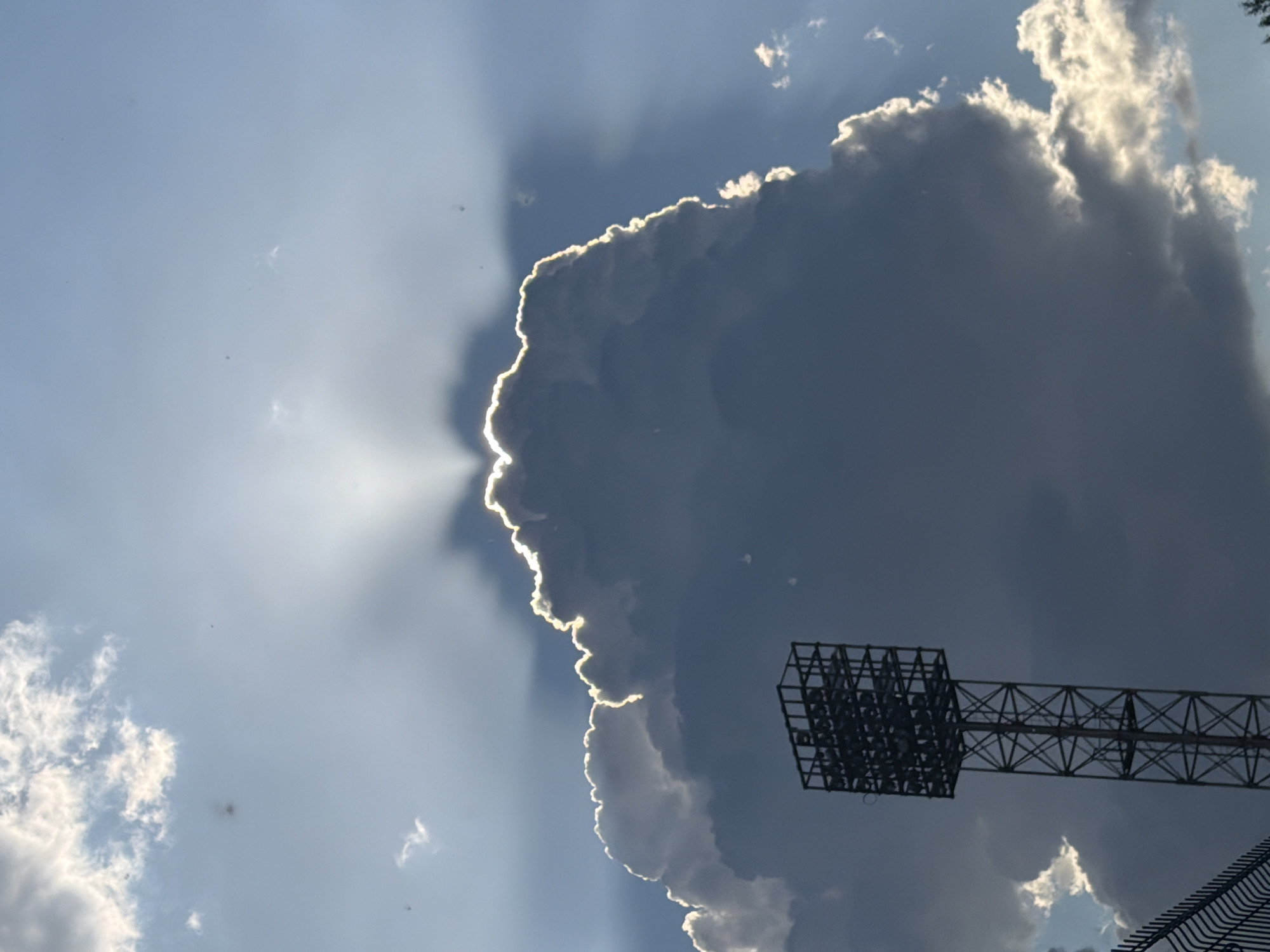 A tall, dark metal floodlight tower stands in the foreground, silhouetted against a dramatic sky. The tower has a square cage-like structure at the top, filled with lights, and is supported by a lattice of intersecting metal beams. To the left and in the lower portion of the image, a dark metal fence is visible. The sky is filled with large, dark clouds that are dramatically illuminated by the sun, which is hidden behind them, creating a halo effect around the cloud edges. Sunbeams are radiating outwards from behind the clouds, creating a celestial display. There are no people visible in the image. The environment appears to be an outdoor sports venue or stadium, based on the presence of the floodlight tower and fence.