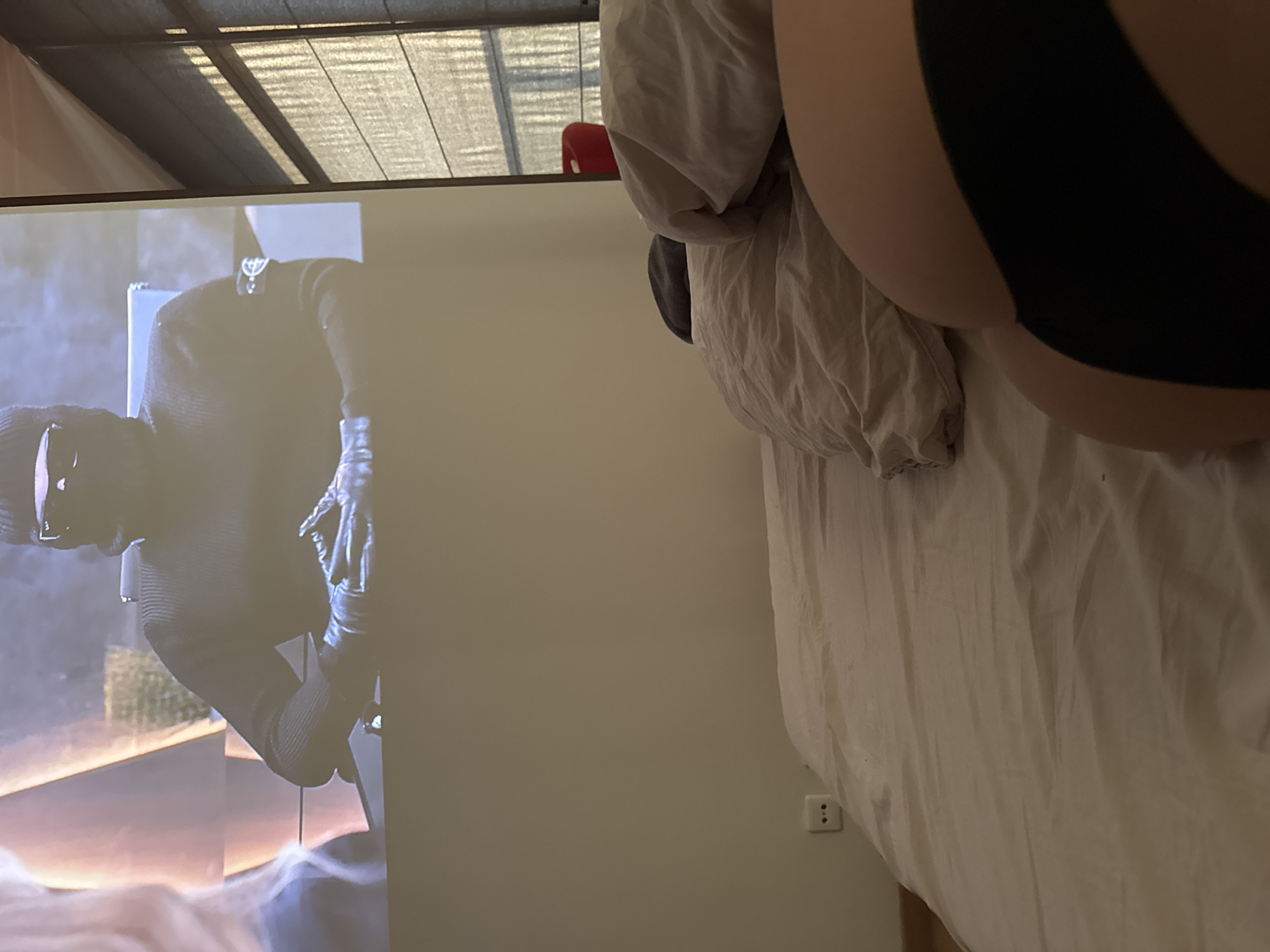 A view from a bedroom features a TV displaying a scene of a person wearing a black balaclava and gloves. The room includes a bed with a duvet and a pillow. An electrical outlet is visible on the wall next to the bed. The location is unknown and cannot be determined from the given image.