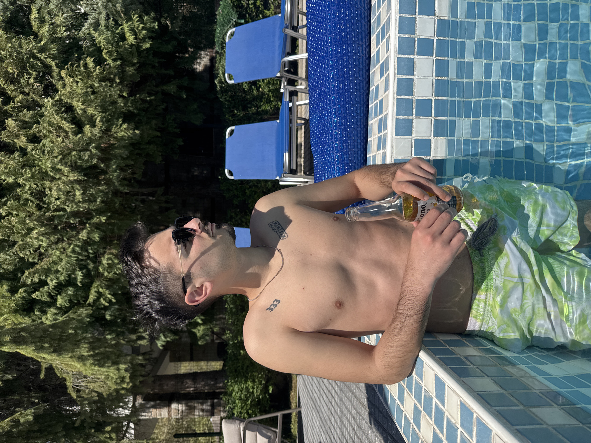 A shirtless man with sunglasses is in a swimming pool, holding a beer bottle. He is wearing green and white patterned swim trunks. The pool is tiled with blue and white squares. Behind him are two blue lawn chairs and some greenery. The location is unspecified and unconfirmed within Marchevo, Bulgaria.
