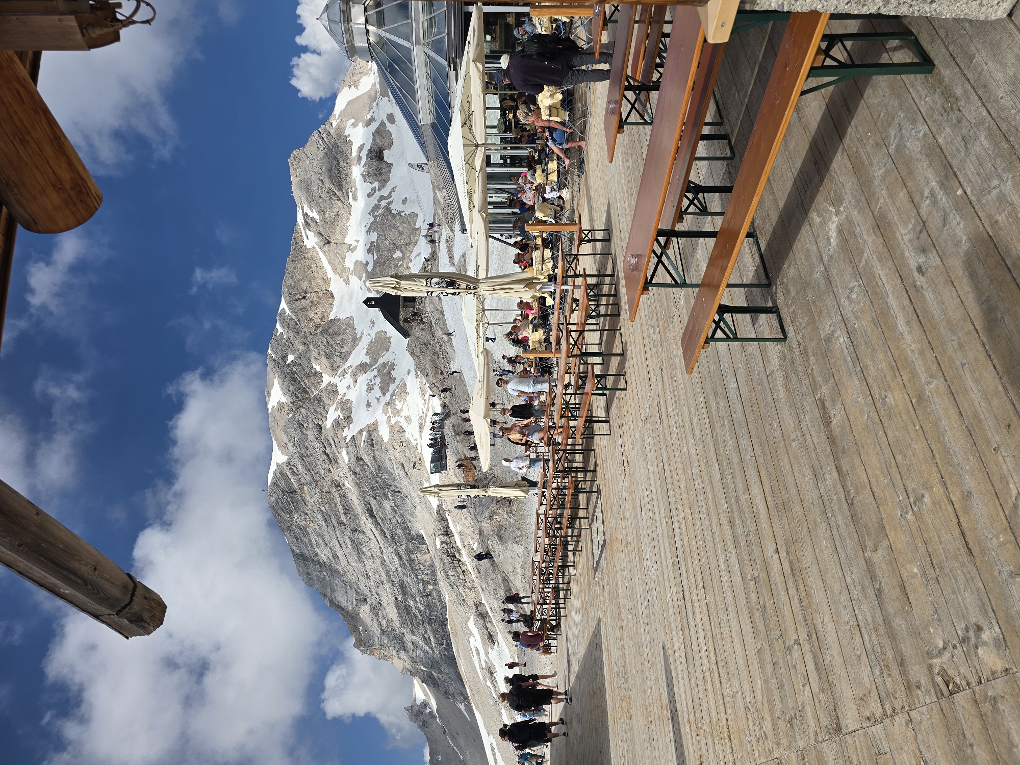 This media file depicts a scenic mountain setting with people enjoying an outdoor dining area. The primary subjects are numerous people casually dressed, seated at long wooden picnic tables and standing in groups. Various objects are visible, including the wooden tables and benches, umbrellas providing shade, and a modern glass-domed building situated on the mountainside. The scene is a spacious wooden deck, suggesting an elevated viewing platform or restaurant terrace.
The activity taking place appears to be a leisurely meal or social gathering on a bright, sunny day. People are interacting, eating, and taking in the mountainous vista. The time of day is likely midday, given the bright sunlight casting distinct shadows. The weather is clear and sunny, with scattered white clouds in a vibrant blue sky.
Notable details include the imposing snow-capped mountain range that forms the backdrop, indicating a high-altitude location. The architecture of the glass-domed building, possibly a visitor center or restaurant, is a prominent feature. The wooden deck itself, with its weathered planks, adds to the rustic yet well-maintained atmosphere. There is no visible text in the image. The overall mood conveyed is one of relaxation and enjoyment of the natural beauty and amenities offered at this mountain resort.