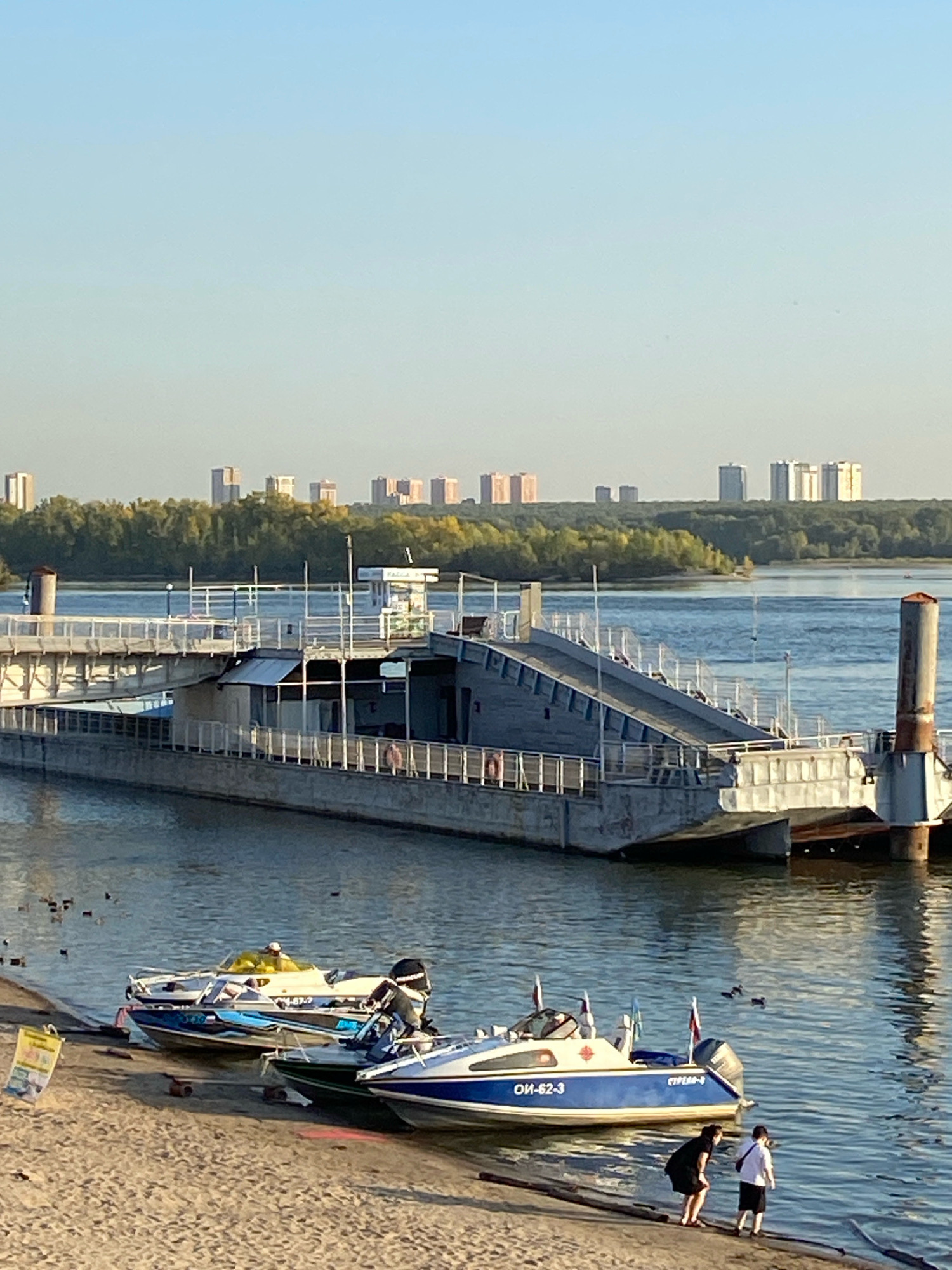 The image shows a wide, calm river with a ferry dock on the right side of the frame and a sandy embankment with boats on the left. In the background, a dense line of trees separates the river from a cityscape of tall, modern buildings under a clear, pale blue sky. The ferry dock is a concrete structure with metal railings and a ramp leading up to a higher level. Several small boats are moored along the sandy bank, with some people standing near them. The water reflects the sky and the structures, creating a tranquil scene. The location is the Ob River in Novosibirsk, Russia, with the city's skyline visible across the water.