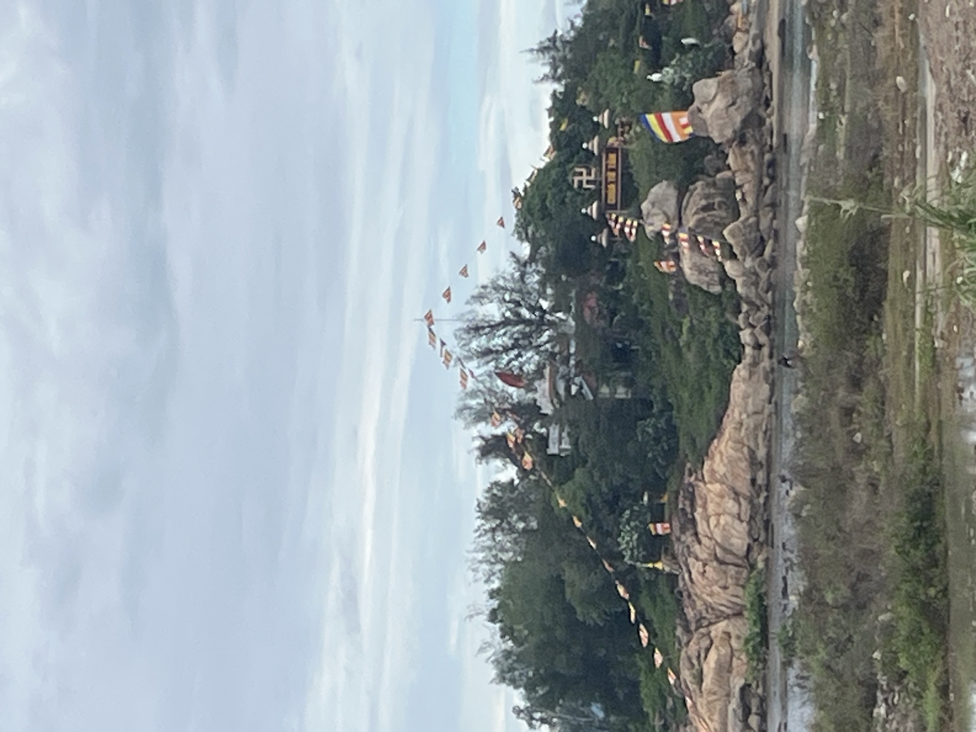 A vertical photograph depicts a hilly landscape in Bac Nha Trang Ward, Vietnam. Prayer flags are strung across the scene. Buildings sit on the slope. The sky is overcast. Vegetation grows on the hill. Boulders are present. A body of water is visible to the right.