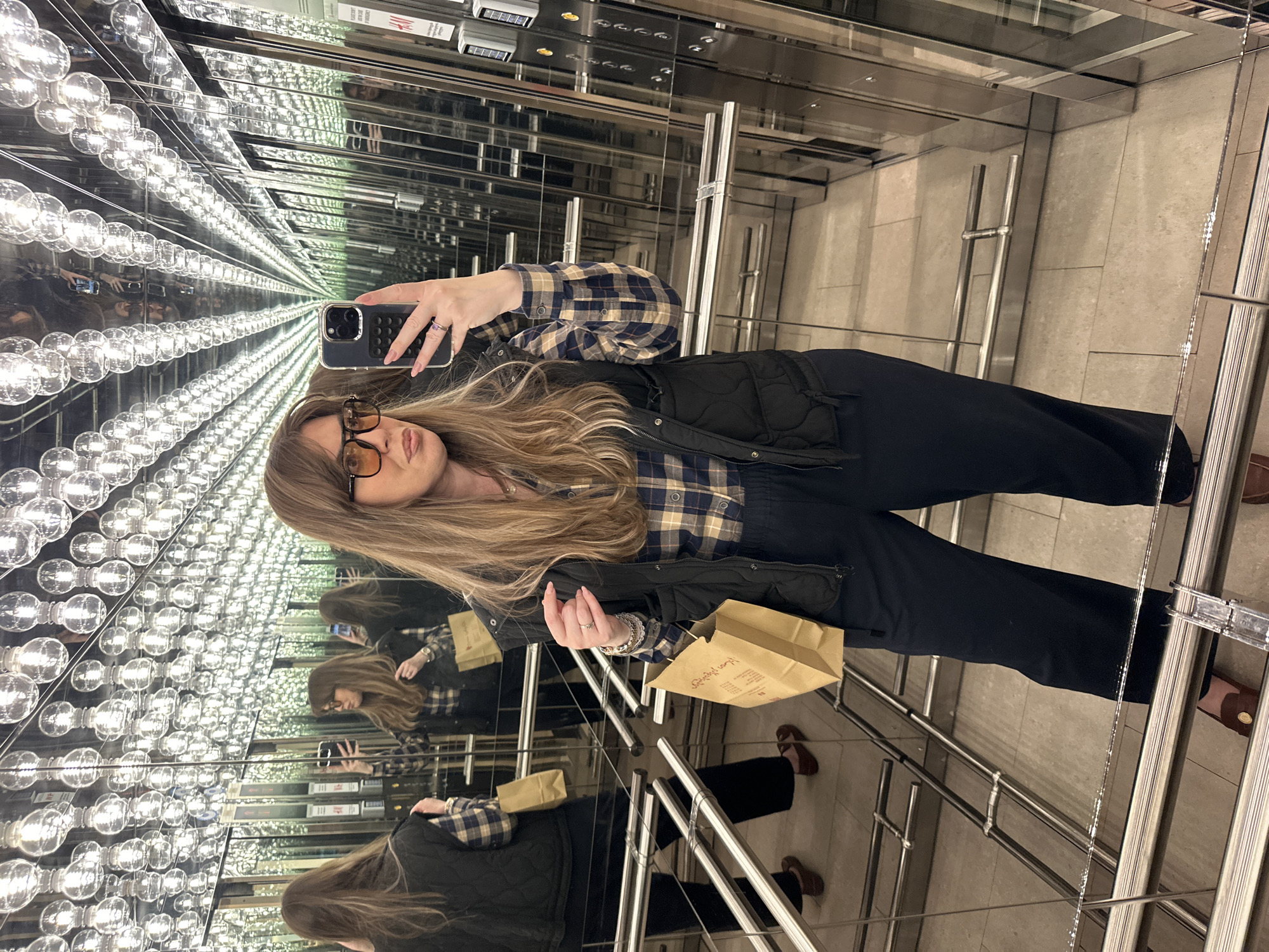 A young woman is taking a selfie in an elevator with walls covered in mirrors and rows of light bulbs, creating an infinity effect. She is wearing glasses, a plaid shirt, a black vest, and dark pants. She holds a smartphone in her right hand and a brown paper bag in her left. The elevator has a gray tiled floor and stainless steel railings. Based on the signage and design details visible, the elevator is likely located in the Galleria Tbilisi shopping mall in Tbilisi, Georgia.