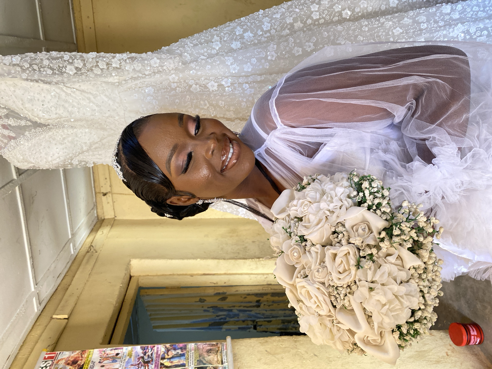 A woman is photographed in Vom, Nigeria. She is wearing a white wedding dress and veil. The dress is ornate with beading and floral embellishments. She is smiling and holding a bouquet of white roses. The environment appears to be a modest building with a painted wall, featuring a doorway with blue trim. There's also a poster near the doorway and a small red and white object (likely a container) near the bouquet.