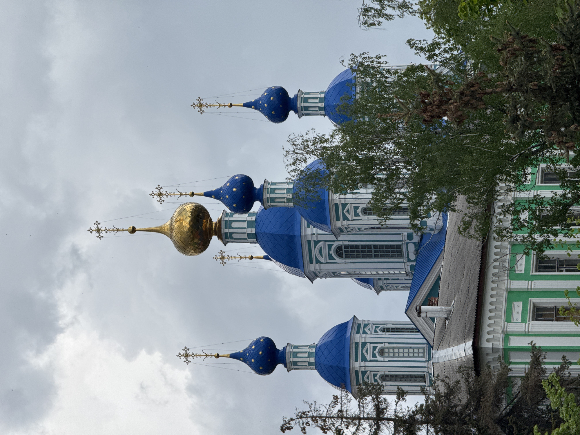 The image captures a building with blue domes, each topped with a golden cross. The building has a green facade with white architectural details around the windows. A tree partially obscures the view of the building, and the sky is overcast. The location is the city of Unknown, Unknown.
