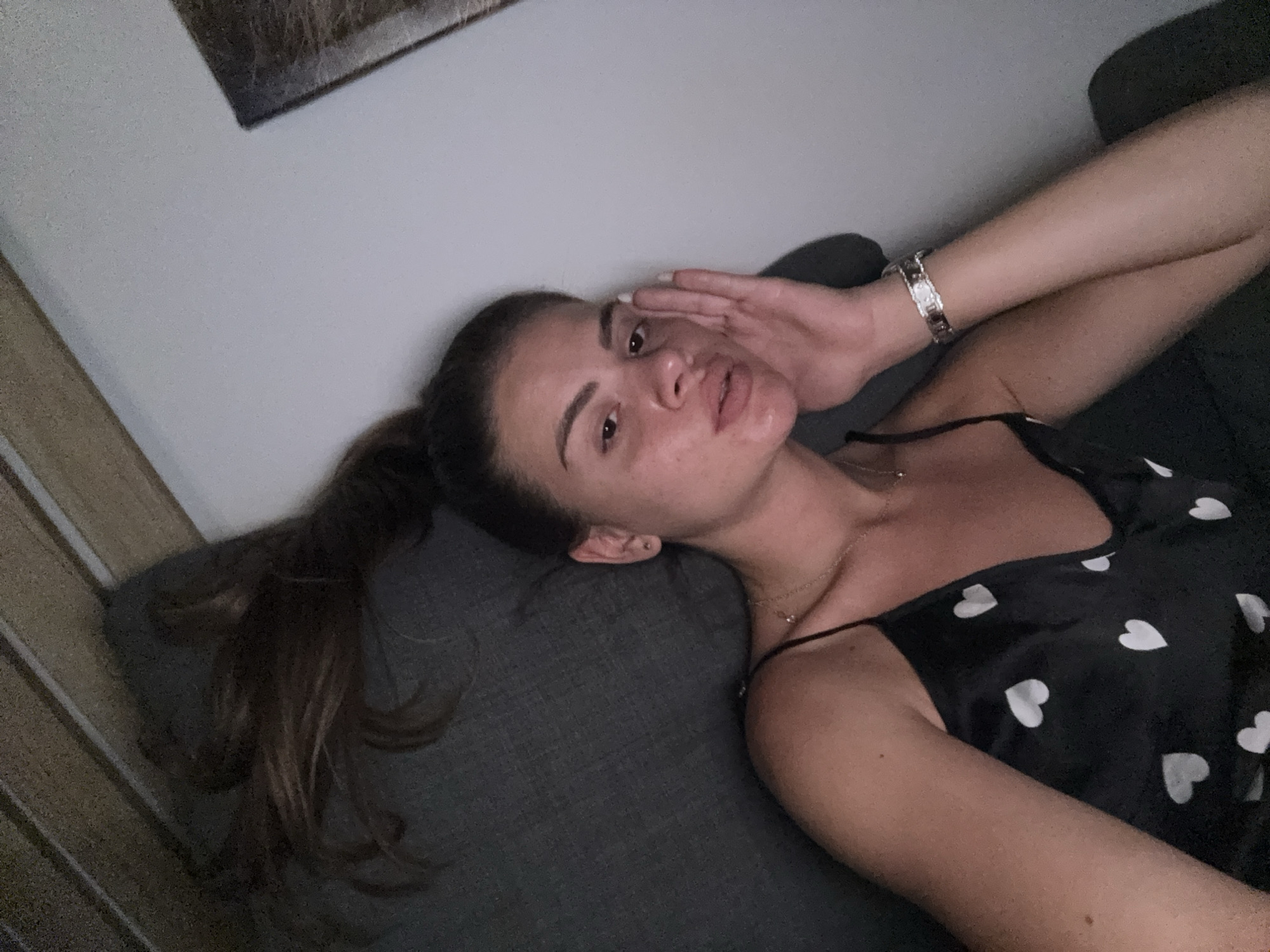 A young woman with brown hair pulled into a ponytail is lying on a dark grey couch. She is wearing a black camisole with white hearts printed on it and a delicate silver necklace. Her right hand is touching her face. A silver bracelet with a white face is on her left wrist. The background shows a light grey wall with a framed artwork hanging on it. The scene is indoors, in what appears to be a living room. The image is taken from a slightly elevated angle, looking down at the woman. There is no information in the image to suggest the location is Plovdiv, Bulgaria.