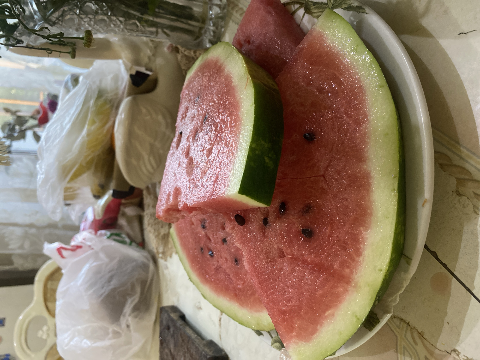 A close-up shot features several slices of ripe watermelon on a white plate. The watermelon slices are vibrant red with visible black seeds and a light green rind. The plate is placed on a table with a patterned surface. In the background, various objects are visible, including plastic bags, a white decorative swan figurine, and a glass vase with wilting flowers. The lighting suggests an indoor setting, possibly a kitchen or dining area. The scene appears to be a still life of food. The location is not specified in the image.