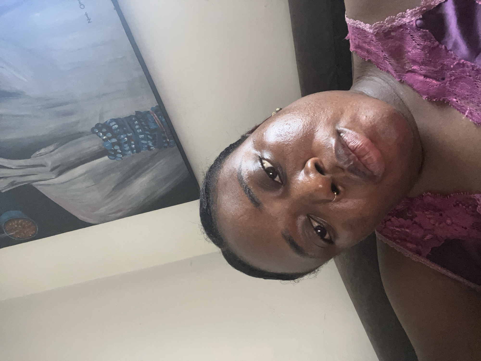 A close-up, portrait-style photograph shows a dark-skinned woman in a purple, lace-trimmed garment. She has a nose ring and dark hair pulled back. Behind her is a painting in a black frame depicting a figure and a container of items. The location is an interior space with white walls. There is no direct indication the location is Abuja, Nigeria.