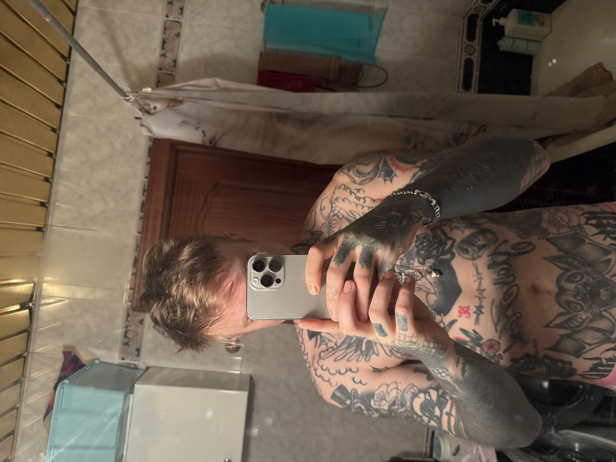 A tattooed individual is taking a selfie in a bathroom. The bathroom features tiled walls and a shower curtain. Various toiletries and other objects are visible on shelves. The individual is holding a smartphone and looking at the screen. The person's tattoos are extensive and cover the arms and torso. The location cannot be confirmed as Moscow, Russia based on the visual information provided.