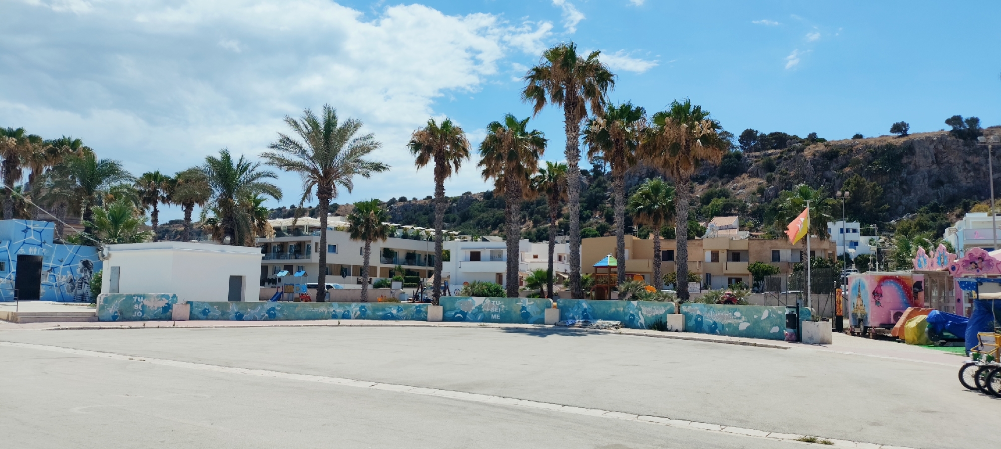 The image depicts a street scene in San Vito Lo Capo, Italy, on a clear, sunny day with some scattered clouds. The foreground is dominated by an asphalt surface, likely a road or parking area, with a curb along its edge. In the mid-ground, several palm trees are prominent, with lush green fronds and textured trunks. Behind the palm trees, there are several buildings, including modern white structures with multiple balconies, and older buildings with a more traditional architectural style. Some buildings appear to be hotels or apartment complexes, suggesting a tourist-oriented area.
To the left of the scene, there's a building with blue-painted walls featuring an abstract mural and a dark doorway. Adjacent to this is a plain white, rectangular building with doorways. A low, blue-painted wall with a decorative pattern runs along the front of these buildings, with some graffiti visible. To the right of the scene, a colorful amusement stall with pink and blue accents is visible, featuring cartoon-like imagery on its sides, including a castle and a rainbow. There are also some children's playground structures visible, including a small slide and climbing frame. A flag, predominantly orange with a yellow section, flies on a pole. In the far background, a rocky hillside covered with sparse vegetation rises, characteristic of the Mediterranean landscape. There are no people clearly visible in the image, and the scene appears to be at rest. The overall impression is a coastal town in a warm climate.