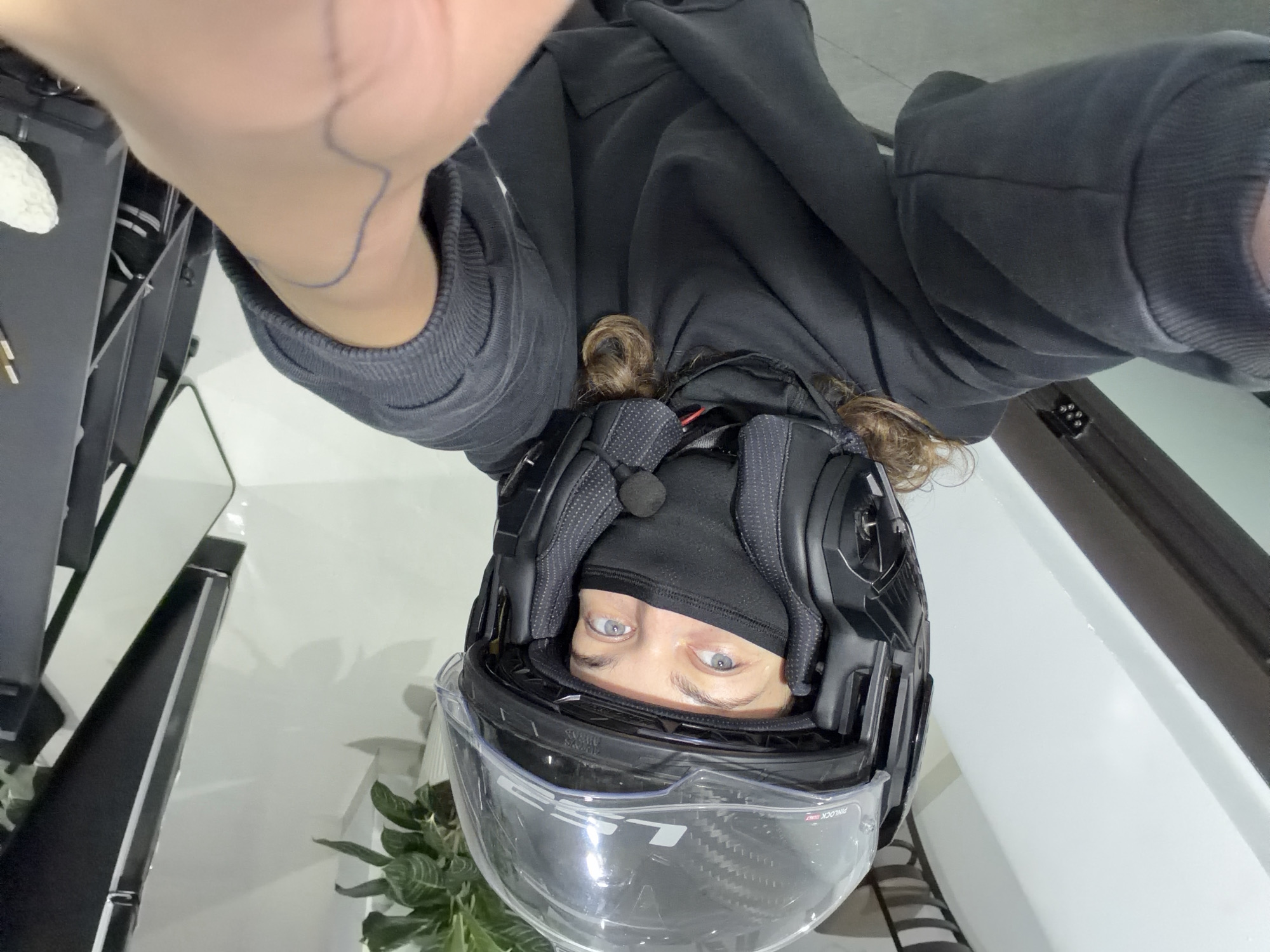 The image is a low-angle selfie, presented rotated 90 degrees counter-clockwise, featuring one person indoors in Đà Nẵng, Vietnam. The subject, a person with striking blue/grey eyes, is seen looking directly into the camera while wearing a black full-face motorcycle helmet, a black balaclava covering most of their face, and a black hooded sweatshirt. Brown, curly hair is visible around the back of the helmet.
The left hand and forearm of the person, holding the camera, are visible at the top of the frame, revealing a faint blue line drawing or tattoo on the wrist area. The black motorcycle helmet has a clear visor, displaying "LS2" and "PINLOCK" branding. A small black microphone is positioned inside the helmet, near the balaclava.
The background consists of bright, clean white surfaces, likely walls or a floor, indicating a modern indoor setting. Dark, angular furniture or structures are visible on the left and right sides of the frame. A green houseplant with striped leaves is partially visible in the lower left, behind the helmet. The activity is the person taking their own photograph, possibly before or after a motorcycle ride, or simply posing indoors. The person's expression appears neutral or intently focused on the camera. The time of day and weather are not discernible from the indoor context.