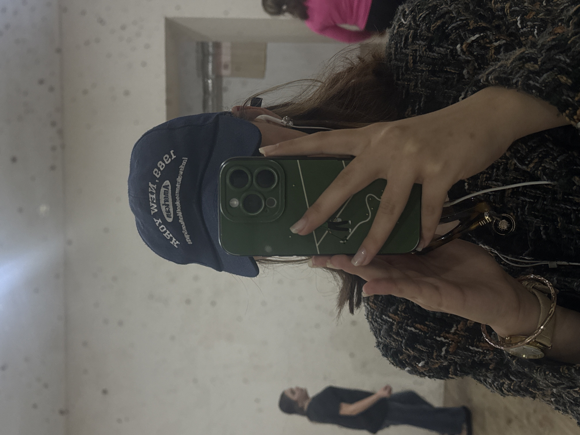 A person is taking a selfie in Yerevan, Armenia. The subject is facing away from the camera in a mirror with a dark blue cap that says "New York" with mirrored text. They are holding a phone with a dark green case with a line drawing. The person is wearing a speckled dark-colored jacket and a watch, with wired headphones in their ear. The selfie captures the reflection of two other people in the background. One person is wearing a bright pink shirt. The other person is walking toward the camera. The room behind the reflection appears to be white with small dark spots.
