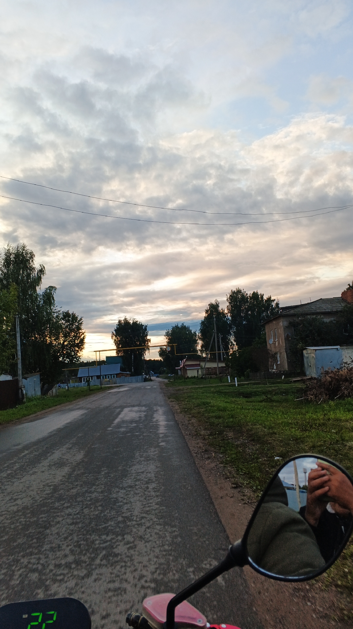 The image captures a scenic view of a village road at dusk, possibly in Podgornoye, Russia, as suggested by the hint. The road, wet and reflective, stretches into the distance, flanked by green grass and trees. Several houses and outbuildings are visible along the roadside, some appearing to be in disrepair. The sky is filled with dramatic clouds, tinged with the warm hues of the setting sun. In the foreground, the reflection in a motorcycle's side mirror shows hands holding a phone, suggesting the photo was taken from a moving vehicle. A digital display showing "22" is also visible in the lower left corner. The overall atmosphere is serene and evokes a sense of rural tranquility.
