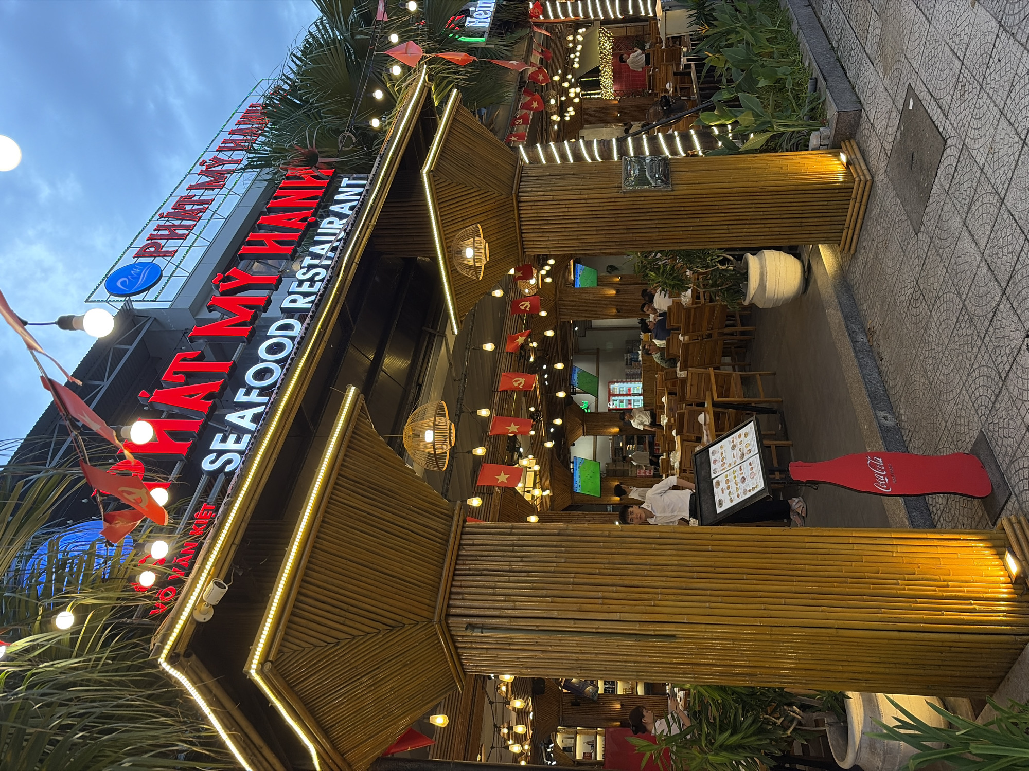 A wide-angle, eye-level, exterior shot captures the entrance of a restaurant in Da Nang, Vietnam, at dusk. The restaurant, named "PHÁT MY HANH SEAFOOD RESTAURANT" as indicated by prominent red neon signage, features a rustic design with bamboo pillars and decorative lighting. Red and yellow flags are strung overhead, adding a festive atmosphere.
Inside, patrons are seated at wooden tables, some engaged in conversation and dining. Several staff members, including a man in a white collared shirt, are visible attending to customers or standing near the entrance. A menu stand is positioned near one of the bamboo pillars, and a red Coca-Cola cutout stands on the tiled pavement.
The environment suggests a busy, welcoming dining establishment. The sky is a muted blue with clouds, indicating the transition from day to night. Lush green foliage surrounds the entrance, contributing to the tropical ambiance. The overall scene depicts a vibrant, illuminated restaurant preparing for or in the midst of an evening service.