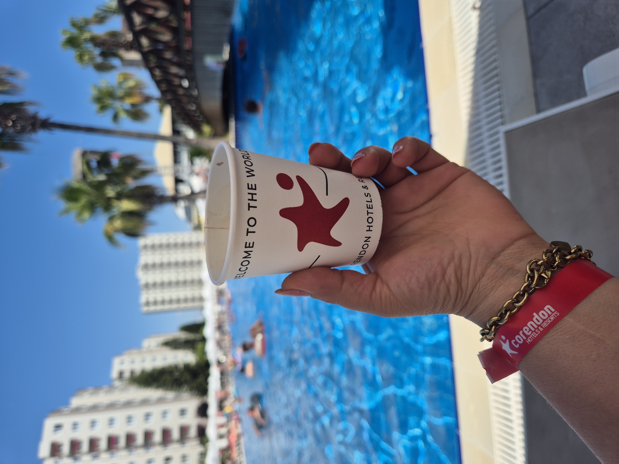 A hand holding a paper cup with a red star logo and "WELCOME TO THE WORLD" printed on it is in the foreground of the image. The hand is also wearing a red wristband with "corendon HOTELS & RESORTS" printed in white and a gold chain bracelet. In the background, a bright blue swimming pool is filled with people relaxing and swimming. Several tall buildings and palm trees are visible beyond the pool. The sky is clear and blue. The scene appears to be a resort or hotel with a pool area.
