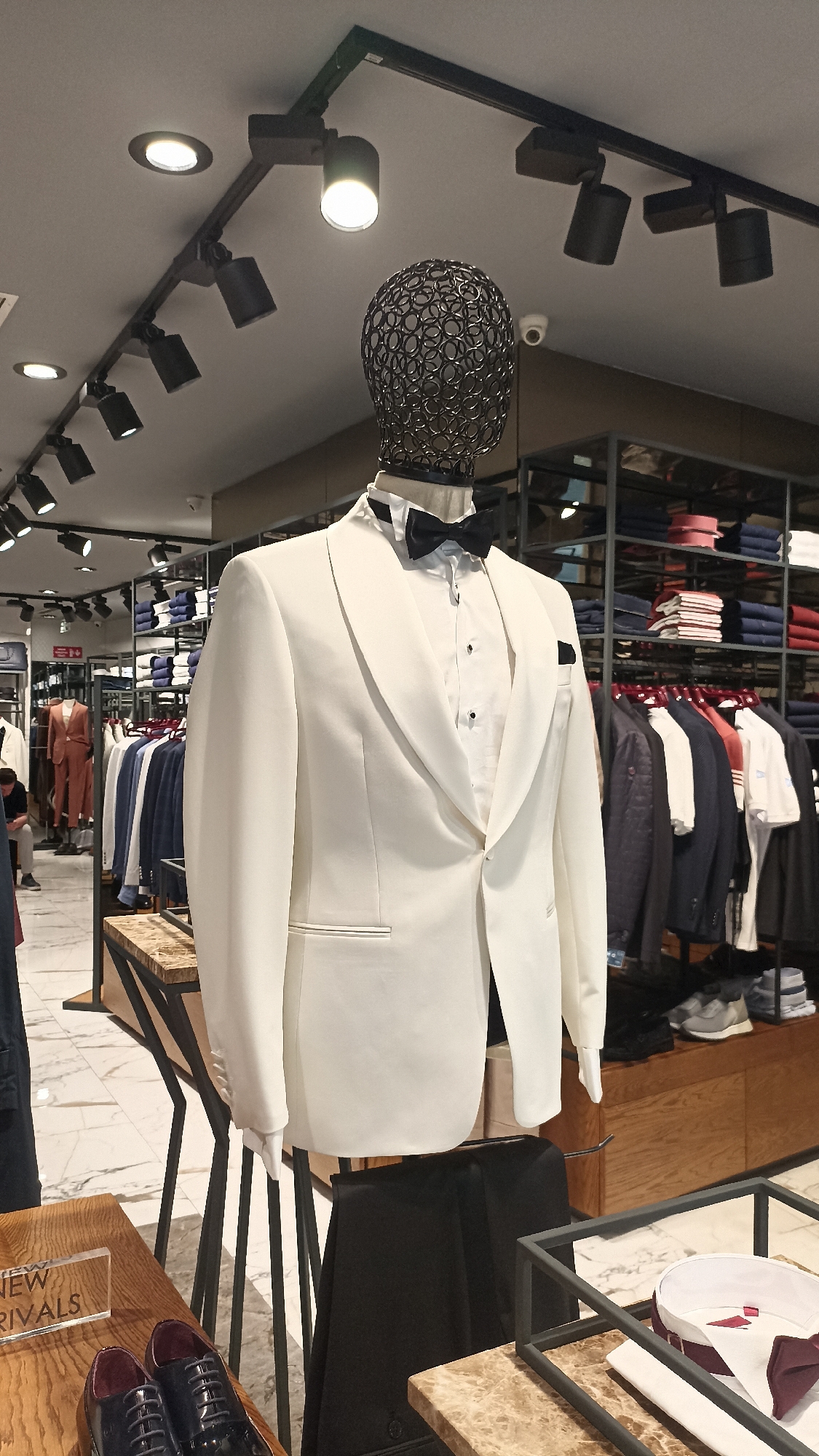 This close-up shot captures a stylish display within a modern men's clothing store in Yenişehir, Turkey. The central focus is a mannequin dressed in a pristine white tuxedo jacket with wide lapels, a white pleated shirt with black stud buttons, and a classic black bow tie. A black pocket square is neatly tucked into the jacket's breast pocket. The mannequin's head is an abstract, black metal mesh design.
The retail environment features track lighting on the ceiling, illuminating the merchandise, and a small, white security camera is visible in the upper right background. Beyond the mannequin, the store is well-stocked with various men's apparel. Racks display jackets and shirts in different colors and styles, while shelves hold neatly folded garments. The flooring is a polished marble with grey veining.
In the mid-ground on the left, a person is seen sitting, and another individual stands further back, suggesting ongoing activity within the store. On a wooden display table in the foreground, a pair of shiny dark formal shoes is presented next to an acrylic block featuring the partially visible text "NEW RIVALS" (likely "NEW ARRIVALS"). Another display stand to the right holds a white shirt with a burgundy tie. The overall impression is that of a contemporary and upscale boutique showcasing formal and casual wear.