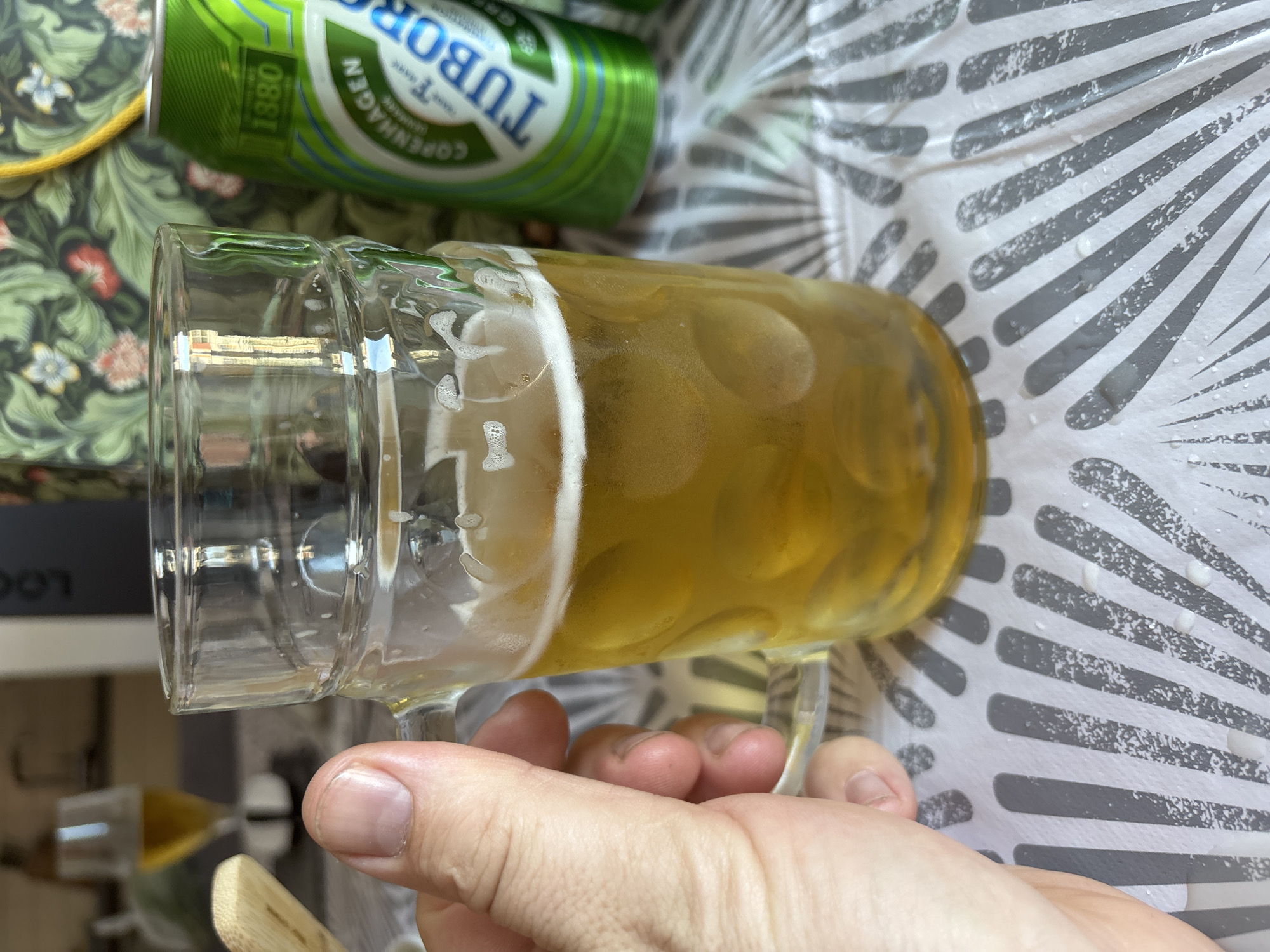 An outdoor close-up shot shows a person's hand holding a beer mug filled with golden lager. The mug is clear glass with a handle and is about two-thirds full. Bubbles are visible on the surface of the beer. Behind the mug, a green can of Tuborg beer is partially visible. In the background, out of focus, is a table with a patterned cloth, and beyond that, a blurred glimpse of a wine glass with a yellowish liquid. The lighting suggests a daytime scene. The image does not provide any information that can place it in the city of Cork, Ireland.
