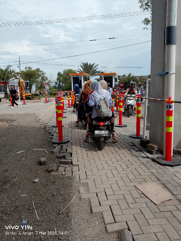 The scene is a road checkpoint with an orange gatehouse in Indramayu, Indonesia, located at coordinates -6.343 latitude and 108.335 longitude. The checkpoint is comprised of a series of orange and yellow striped poles with a yellow and black metal barrier. There are several motorcycles lined up in front of the gatehouse. A man in a white shirt and blue jeans is walking towards the gatehouse, while a group of women wearing headscarves are standing behind their motorcycles waiting. The scene is observed from the perspective of someone standing to the right of the checkpoint. The road surface is made of gray concrete blocks, with some of them missing, leaving loose gravel in their place. There is a yellow and black sign at the checkpoint that reads "BEL". The motorcycles are parked in a single file line, with the motorcycles in the back of the line blocking the view of the individuals behind the motorcycles. The gatehouse is made of yellow, orange, and black materials. There is a bright blue sky with a few thin clouds above the gatehouse. The image was taken with a phone camera. The timestamp on the image reads “7 Mar 2025, 14:01”. The camera model is “vivo Y19s.” There are power lines that run across the top of the image.