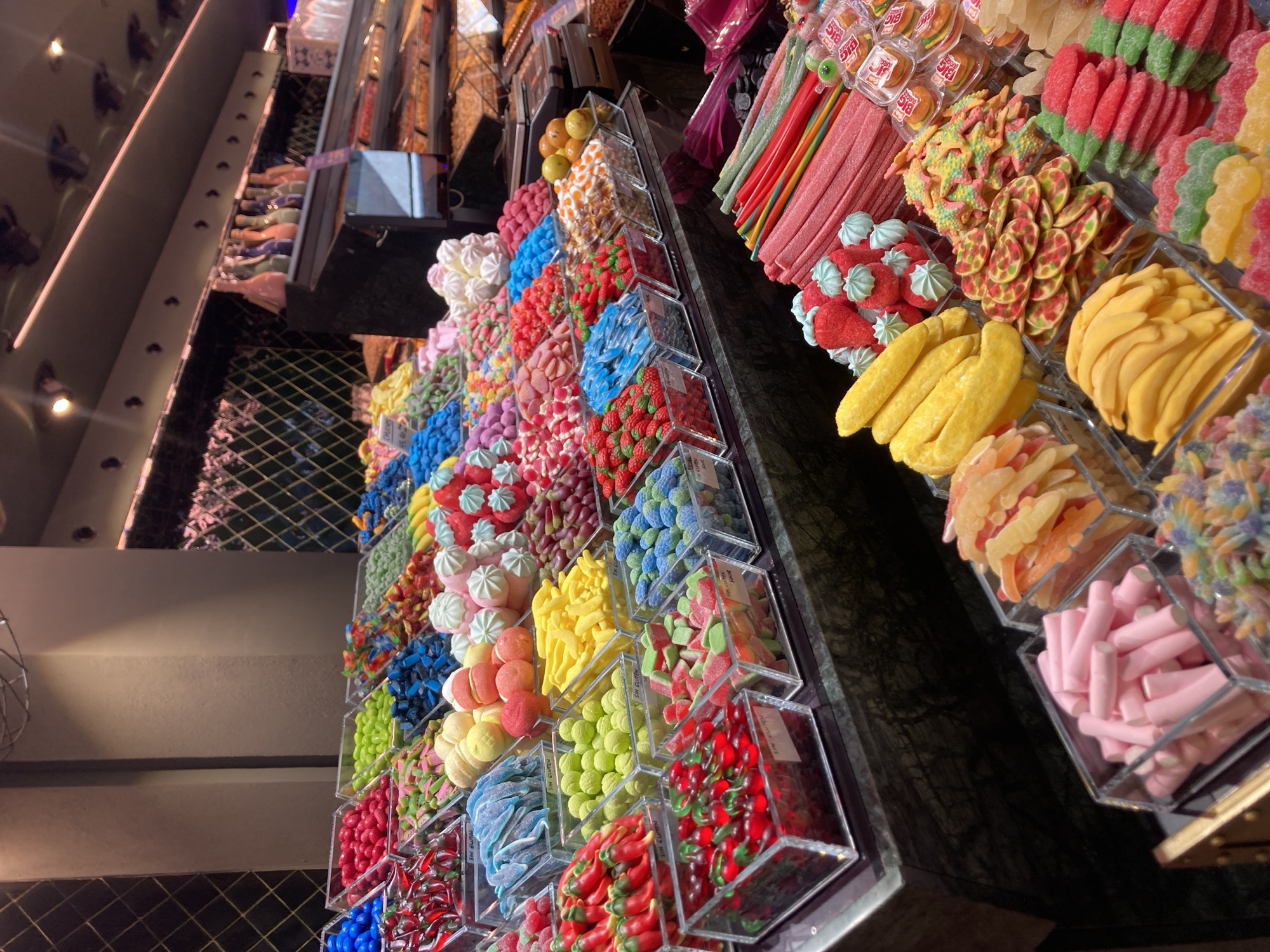 The image shows a vibrant display of assorted candies in a store, likely a confectionary or a section of a market. The candies are arranged in clear plastic containers, showcasing a wide variety of colors, shapes, and textures. Some of the visible candies include gummy bears, sour belts, fruit-shaped gummies, marshmallows, and what appear to be licorice sticks. In the background, more rows of candies are visible, as well as shelves stocked with other products, some of which appear to be packaged goods. The lighting suggests an indoor environment, possibly a brightly lit retail space. The overall impression is one of abundance and a sensory overload of sweet treats. This scene is consistent with the offerings found in many shops in the La Boqueria Market on Las Ramblas in Barcelona, Spain, which is renowned for its diverse food stalls, including those selling a wide array of candies.