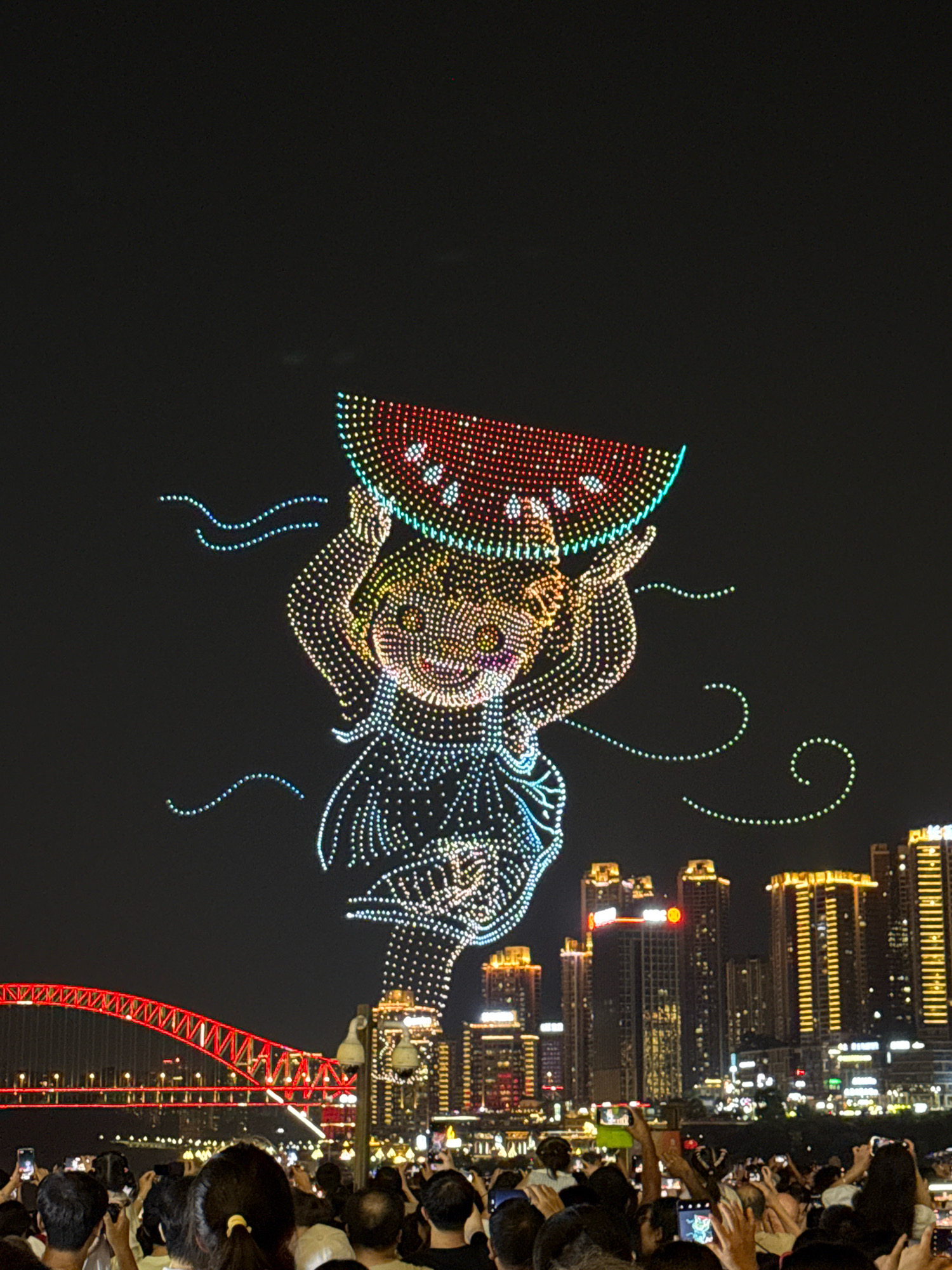 An aerial drone light show is in progress over a cityscape at night. The main subject is a drone formation creating the image of a person holding a slice of watermelon. The figure is composed of numerous small, colored lights, predominantly yellow and blue, with red and white lights forming the watermelon. The watermelon slice is depicted with red seeds. To the left of the figure, several blue dotted lines curve in the air, resembling wisps of smoke or water. In the background, a city skyline is visible, featuring several illuminated skyscrapers with golden-yellow lights. A prominent red-lit bridge spans across a body of water in the middle ground. In the foreground, a crowd of people are gathered, many holding up their mobile phones to capture the spectacle. The overall environment is dark, indicating nighttime, with the lights from the drones and city providing illumination. The scene suggests a public event or celebration.