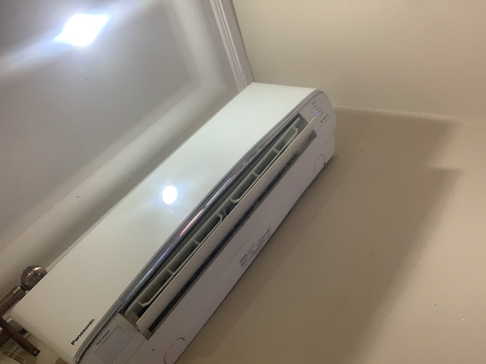 The image shows a close-up, low-angle view of a white Panasonic air conditioning unit mounted on a light brown wall. The unit is angled upwards and to the right, with its louvers partially visible. The "Panasonic" logo is printed in black on the upper left side of the unit. A bright light source reflects off the ceiling, which has crown molding. The scene appears to be indoors, possibly in a residential or commercial setting in Abuja, Nigeria. There are no people or actions depicted in the image.