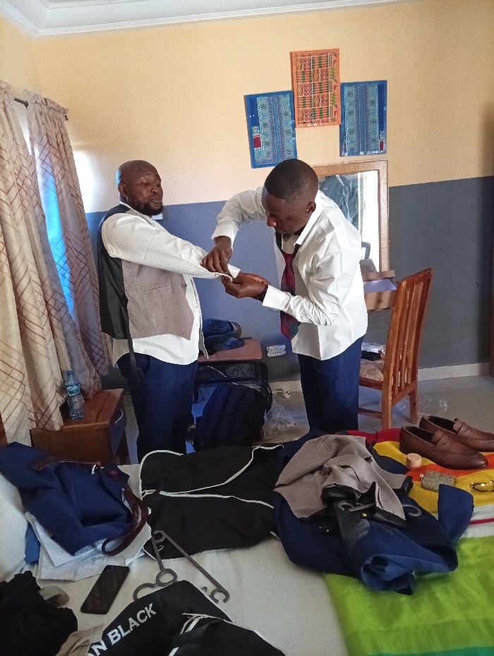 Two men are in a room. One helps the other with his cufflink. The first man wears a white shirt, gray vest, and dark pants. The second man wears a white shirt with a red tie and dark pants. A bed is covered in clothing and accessories, including a black and white shirt that says "IRON BLACK." Two brown shoes are on a colorful striped cloth. Luggage sits behind the two men. A wooden chair is positioned to the right of the men, near a mirror.