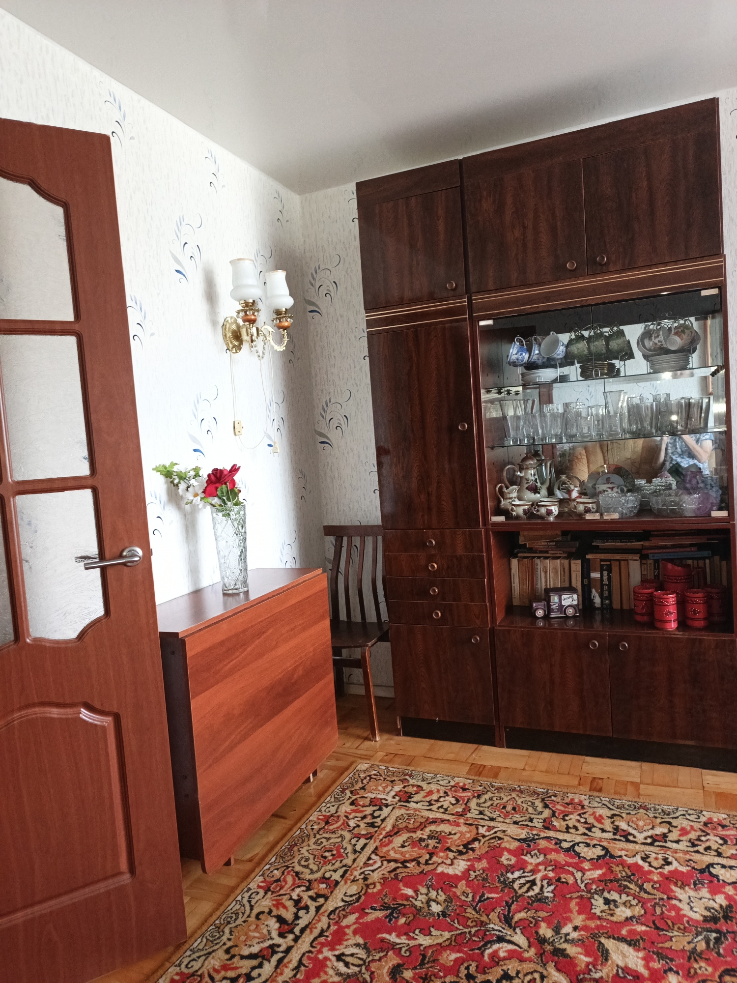 The image shows an interior scene that appears to be a living room or dining room. A wooden door with glass panes is on the left side of the frame. To the right of the door is a wooden folding table with a vase of flowers on it. Further to the right is a large, dark wooden cabinet with glass-fronted shelves displaying various dishes, glassware, and decorative items. Below the glass shelves are wooden drawers and cabinets. A wooden chair is partially visible to the right of the table. A large, patterned rug covers the wooden floor in the foreground. The walls are covered with light-colored wallpaper with a subtle blue and grey floral pattern. A wall-mounted light fixture with two lamps is on the wall between the door and the cabinet.
This image is not located in the city of Kazan, Russia.