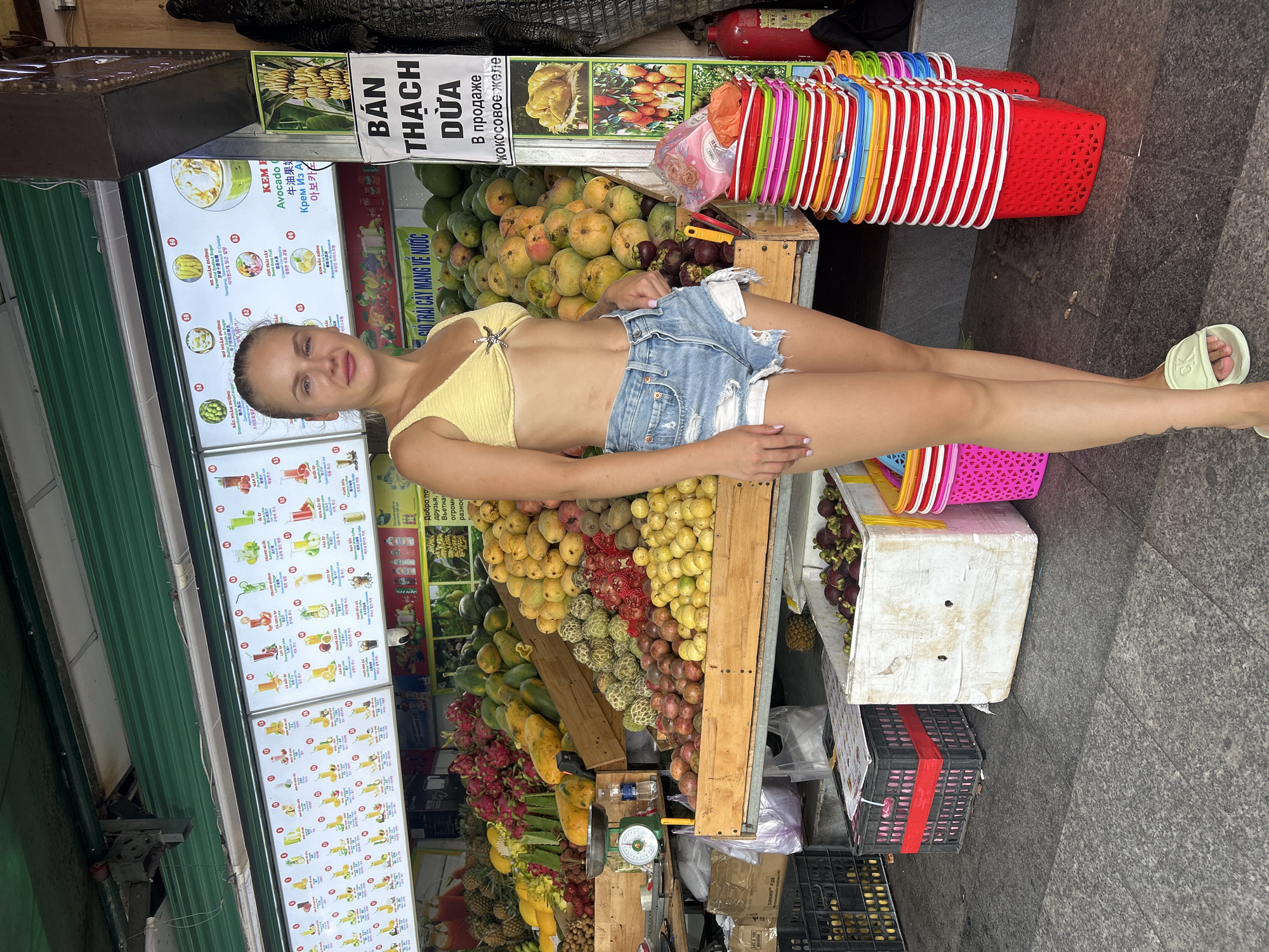 A woman stands in front of a fruit stand in Nha Trang, Vietnam. She is wearing a yellow bikini top and denim shorts. The fruit stand is filled with a variety of fruits, including mangoes, pineapples, and dragon fruit. There are also menus displayed above the fruit stand, showing different types of drinks. In the background, a large statue of an alligator is visible. The ground is paved with granite tiles.