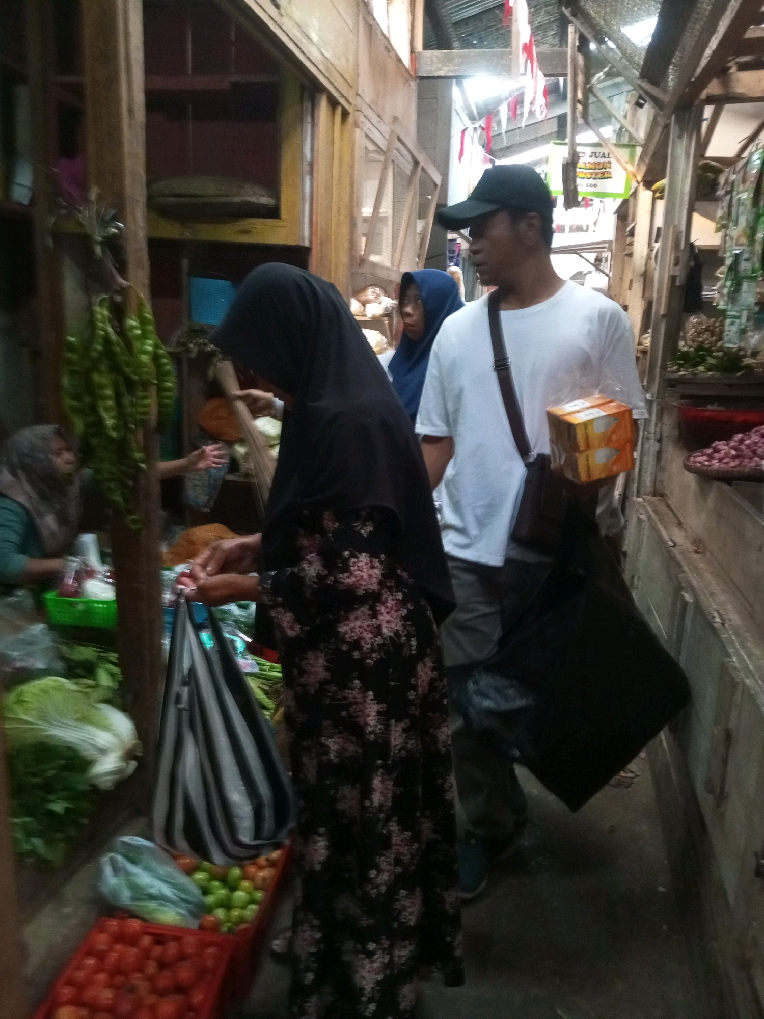 This image depicts a market scene in Kalibeber, Indonesia. In the foreground, a woman wearing a black hijab and a floral dress is holding a bunch of green vegetables. Beside her, a striped bag hangs from a stall. To her right, a man in a white t-shirt and a black baseball cap is holding two boxes of tissues. He has a dark bag slung over his shoulder. Behind them, other market stalls are visible, with various produce and goods displayed. In the background, red and white banners are hanging, suggesting a festive occasion. The market appears to be open-air, with wooden structures and corrugated metal roofing. Several other individuals are present in the market, including a woman in a blue hijab in the middle ground. The overall atmosphere suggests a busy, everyday market environment.