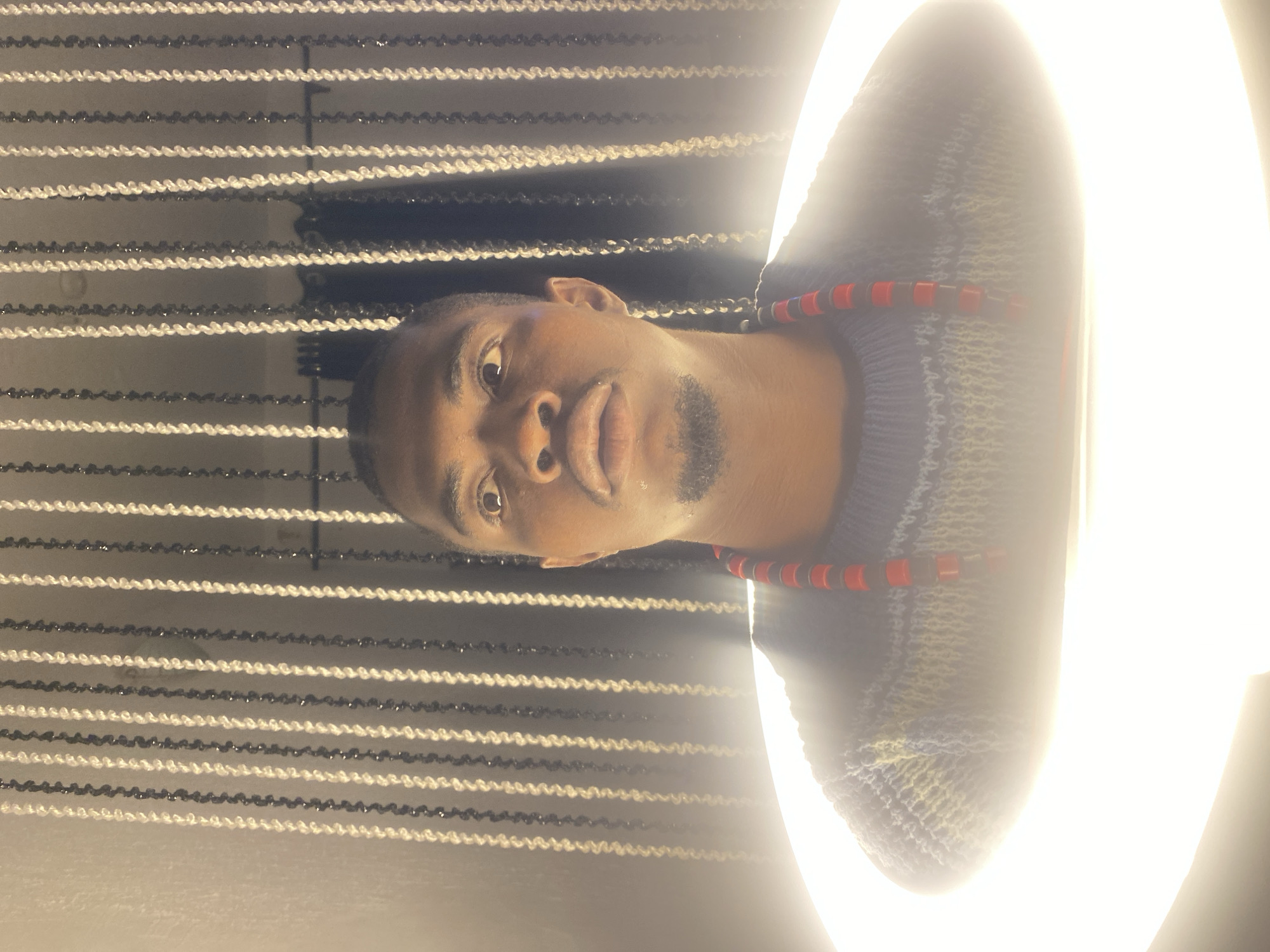 A medium shot captures a dark-skinned man from the chest up, looking directly at the camera with wide eyes. He has a light beard and is wearing a gray, blue, and yellow patterned sweater. A red beaded necklace adorns his neck. He is positioned within a bright, circular light source, likely a ring light, which obscures the lower portion of his body and casts a strong glow. Behind him, a curtain of vertical strands, alternating between black and white beads, hangs against a plain wall. The overall lighting is dramatic, with the ring light creating a halo effect around the man. The image appears to be a self-portrait or a posed photograph taken indoors. There is no identifiable landmark or detail to indicate the specific location within the city of Abakaliki, Nigeria.