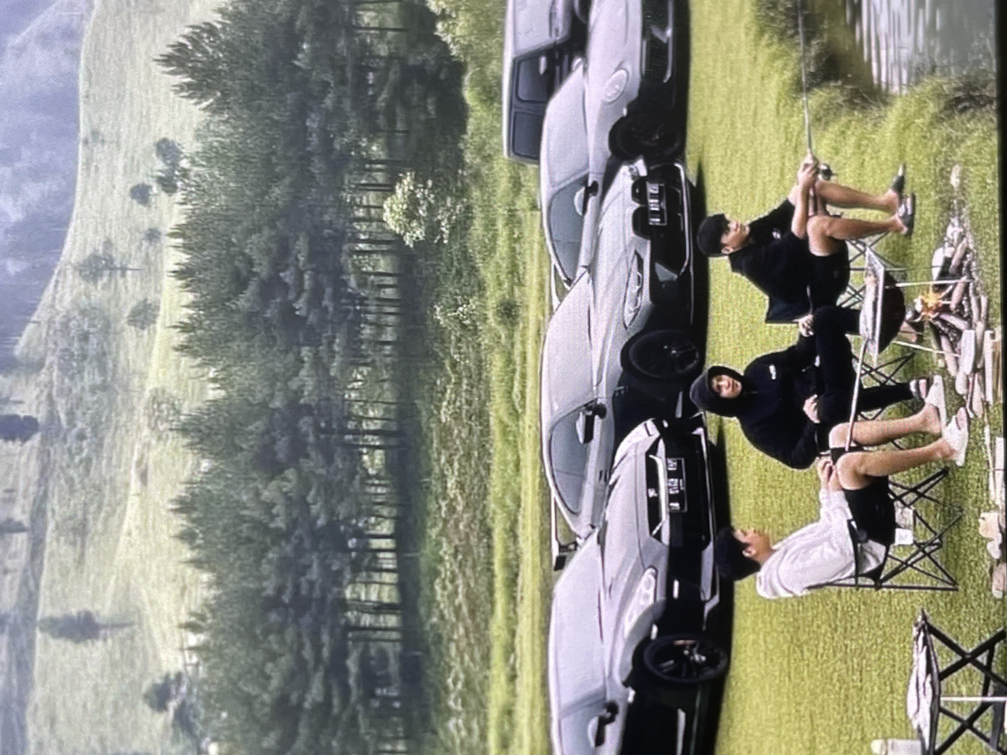 In an outdoor setting, three young men are present. Two of them are seated on folding chairs, with one holding a fishing rod that extends towards the water. A third individual is also seated. In the background, several luxury cars are parked on a grassy area. The environment appears to be a natural landscape with trees and a body of water. There is also a small campfire with cooking equipment visible.