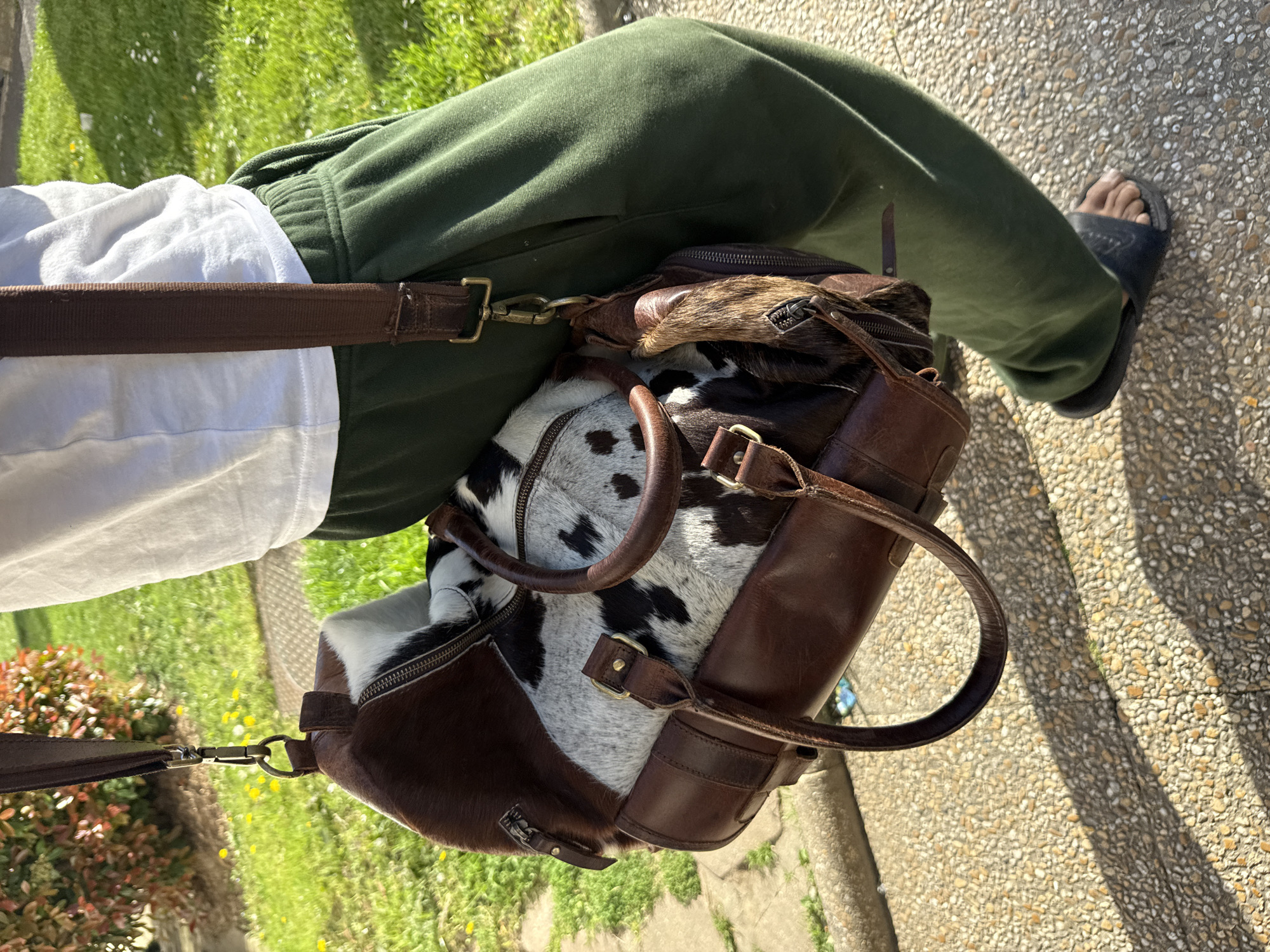A person, presumably sitting, has their left leg (clad in green sweatpants) visible on the right side of the image and a partially visible right leg is seen to the left. A white t-shirt covers the upper torso. A leather and cowhide bag sits on the person's lap. The bag is primarily brown leather with patches of black and white cowhide. The bag has two brown leather handles and a shoulder strap with a silver-colored clip attached. The person is wearing black sandals. The location appears to be outdoors, with grass and flowers visible in the background. The ground is paved with small stones. The image orientation is a 90 degree clockwise rotation of the original. There is no evidence to suggest that the image was taken in Sarcelles, France.