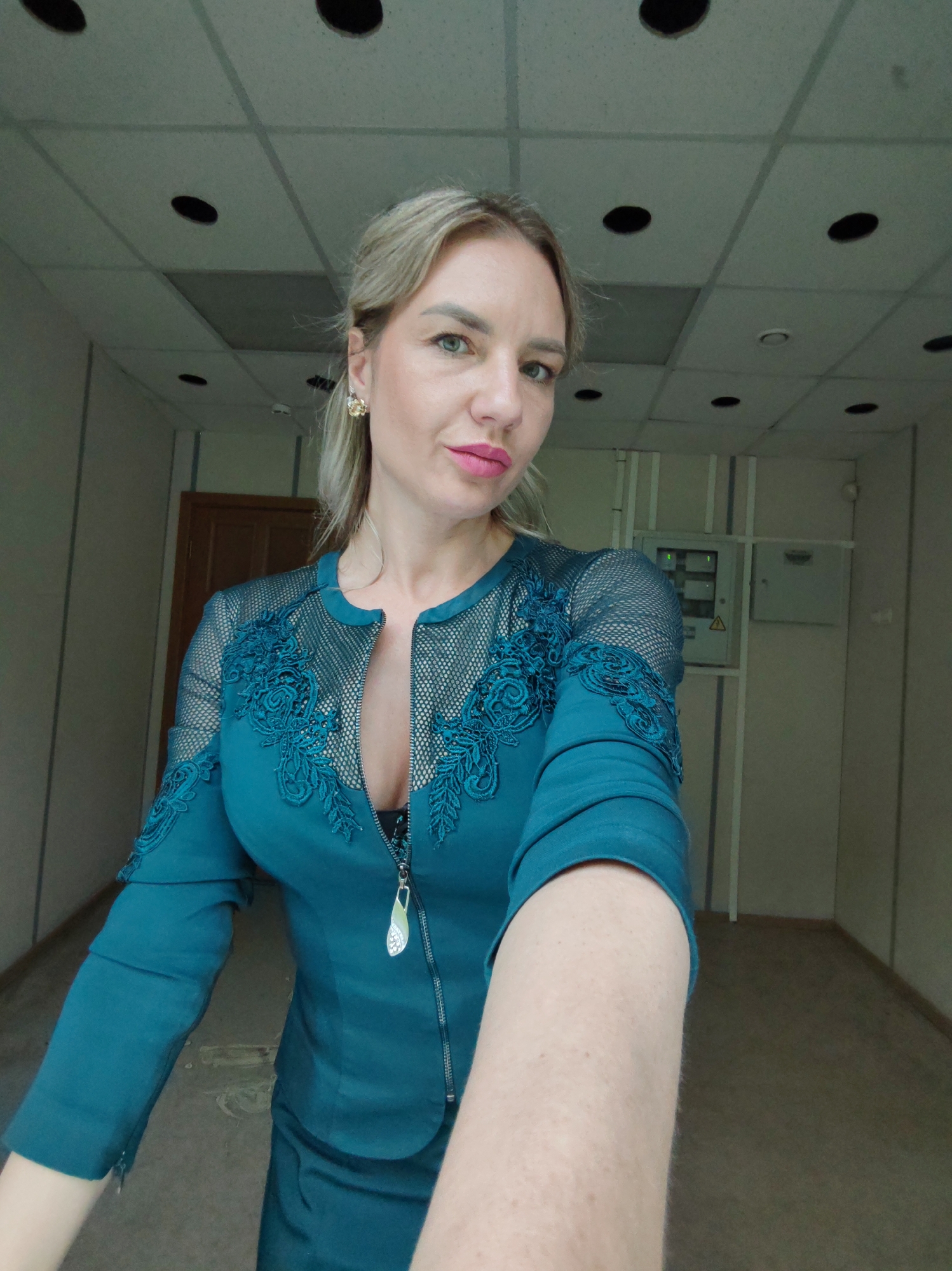 A woman with blonde hair pulled back and wearing a teal dress with lace and mesh accents stands in what appears to be a vacant office or hallway. She is taking a selfie, with her right arm extended towards the camera. Her makeup includes pink lipstick and eyeliner. She is wearing a gold earring in her left ear. The background shows a dropped ceiling with circular light fixtures, beige walls, and a wooden door. There is also an electrical panel visible on the wall. The floor is a neutral color and appears to be carpeted.