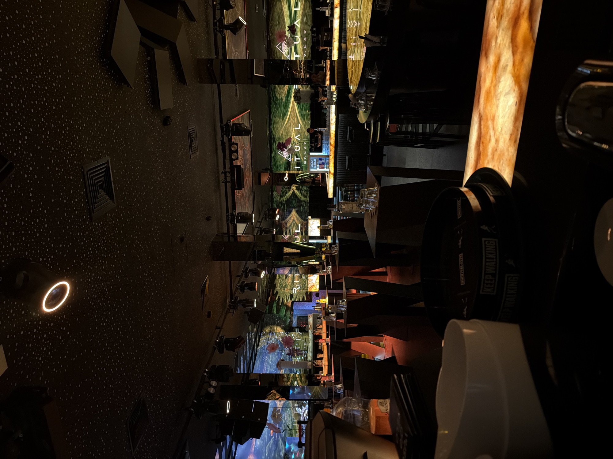 The image shows the interior of "RITUAL BY LAVI," a bar/restaurant in Sofia, Bulgaria. The camera is positioned near the bar, with a partial view of a bar counter and a "Keep Walking" branded container. The bar area is dimly lit with accent lighting. Digital projections with the "RITUAL BY LAVI" logo are visible on the back wall, creating a dynamic visual environment. The foreground is dominated by objects related to service or consumption in the establishment.