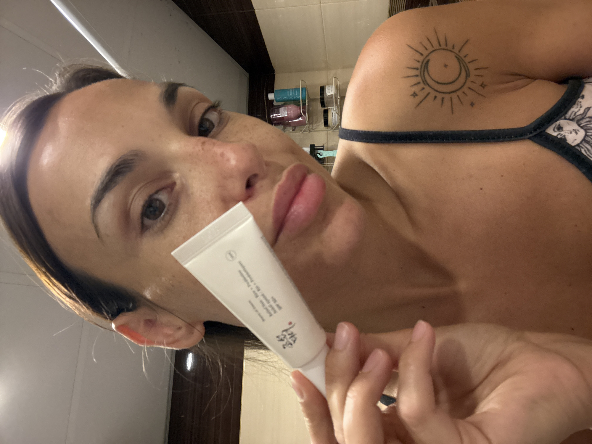 A close-up, over-the-shoulder selfie of a woman holding a tube of skincare product. She is looking towards the camera with a neutral expression. She has light brown hair pulled back, and freckles on her face. She is wearing a dark-colored tank top, and has a tattoo of a sun and moon on her left shoulder. In the background, a shelf with various bottles and jars is visible. The environment appears to be a bathroom, with tiled walls. The location of this image is not discernible from the visual information provided.
