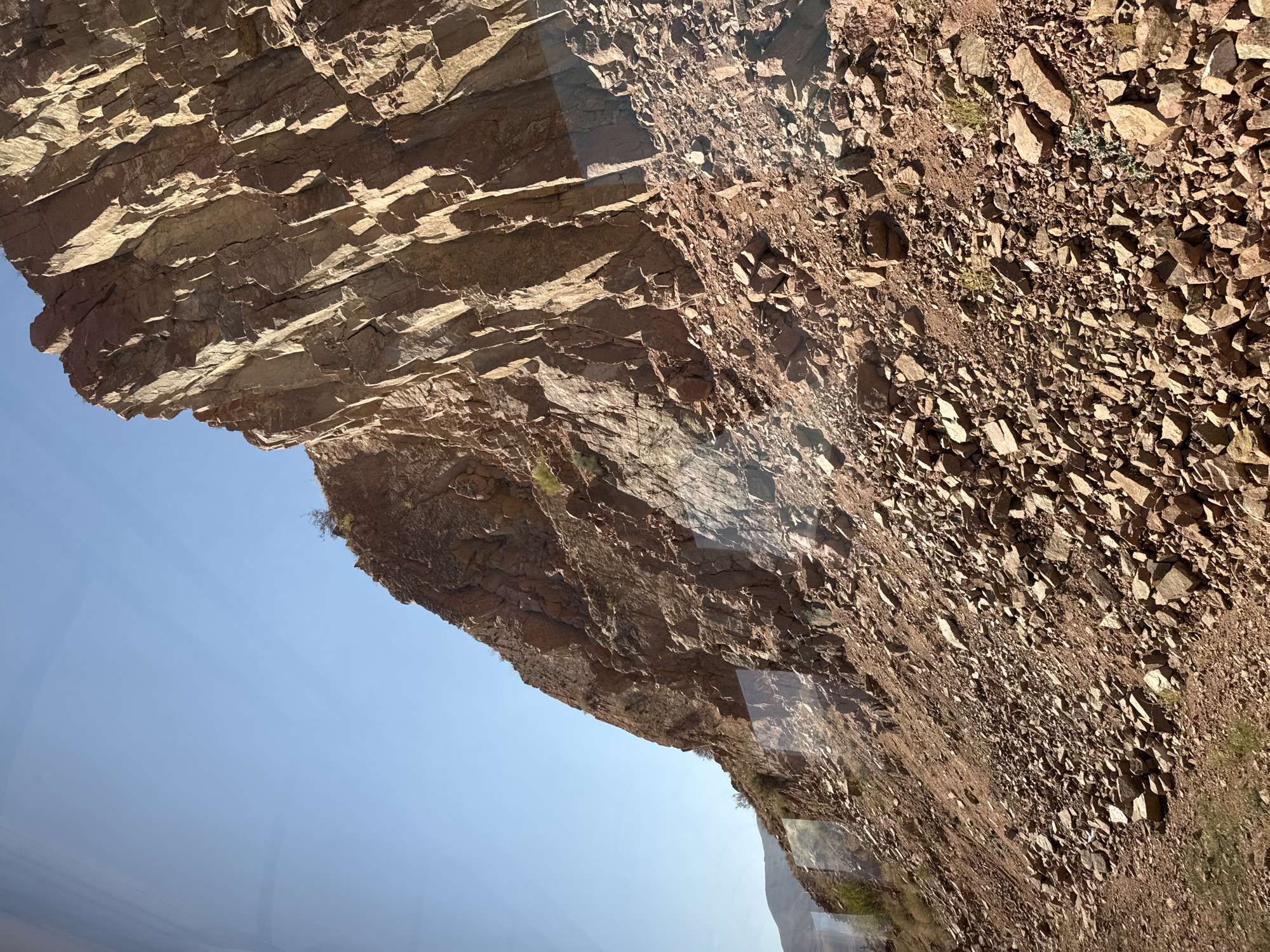 This image depicts a natural scene of a steep, rocky mountainside under a clear blue sky. The terrain is characterized by loose, angular rocks and boulders in shades of reddish-brown, covering a significant portion of the frame. Large rock formations with distinct vertical striations rise sharply on the right side of the image, casting shadows that highlight their textured surfaces. Sparse, dry vegetation is visible clinging to the slopes. The foreground shows more of the rocky debris. Reflections are present on a transparent surface, suggesting the photo was taken through a window. The scene does not contain any people or man-made structures, and no specific landmarks are identifiable to pinpoint a location within Sasyk-Bulak, Kyrgyzstan.