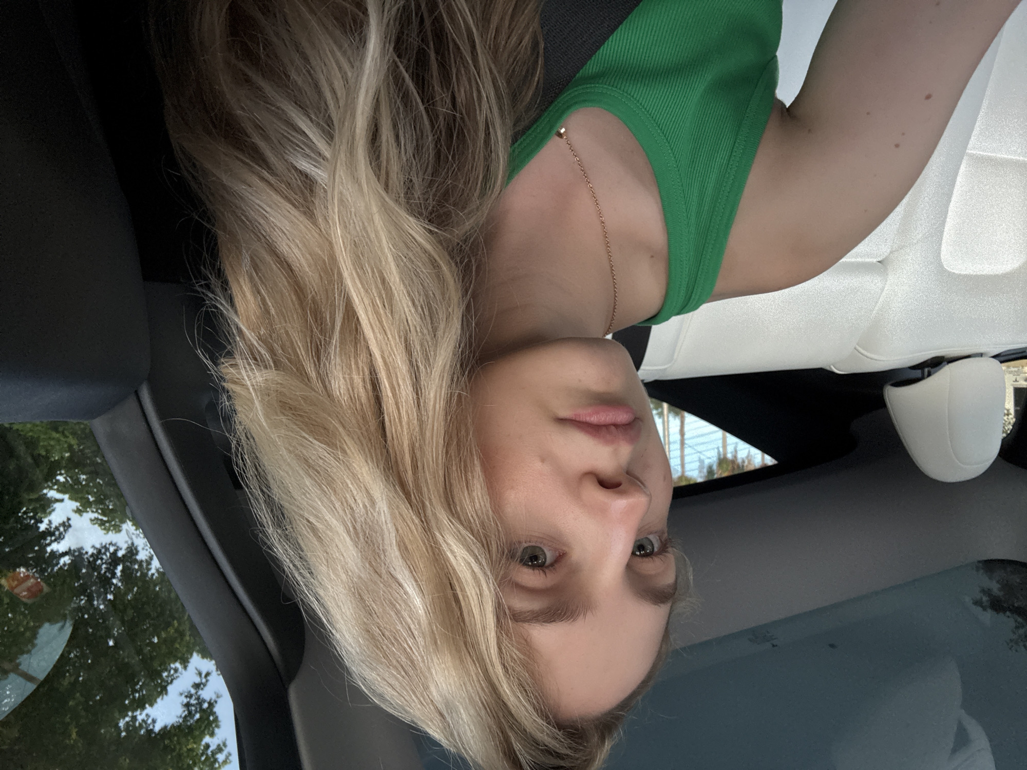 A young Caucasian woman with long, wavy blonde hair is shown from the chest up, seated in the passenger seat of a vehicle. She is wearing a green tank top and a rose gold necklace with a small pendant. The interior of the car is visible, including white leather seats and a dark-colored dashboard and pillar. The car has a panoramic glass roof, through which the sky and trees are visible. The scene appears to be in a suburban or residential area, with lush green trees and a cloudy sky suggesting an outdoor setting. The woman is looking slightly away from the camera with a neutral expression.