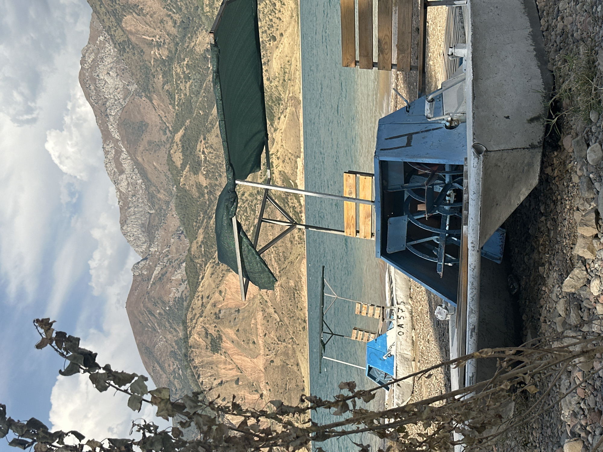A bright, sunny day at a lake in Yusuphana, Uzbekistan, features a shoreline with several blue and white paddle boats lined up on a gravelly beach. In the foreground, a paddle boat is prominently positioned, showing its concrete hull, blue metal casing with pedal-driven wheels, and wooden seating. Behind it, another paddle boat with a blue front and a green canopy is visible. Further back, a third paddle boat with a blue front and a wooden bench is partially obscured. The lake itself is a vibrant blue, with ripples on its surface. The backdrop consists of imposing, rugged mountains with a mixture of rocky and vegetated slopes, under a sky with scattered white clouds. A dry, leafy branch extends into the frame from the left, adding a touch of natural detail. The scene is tranquil and appears to be a popular spot for recreational activities by the water.