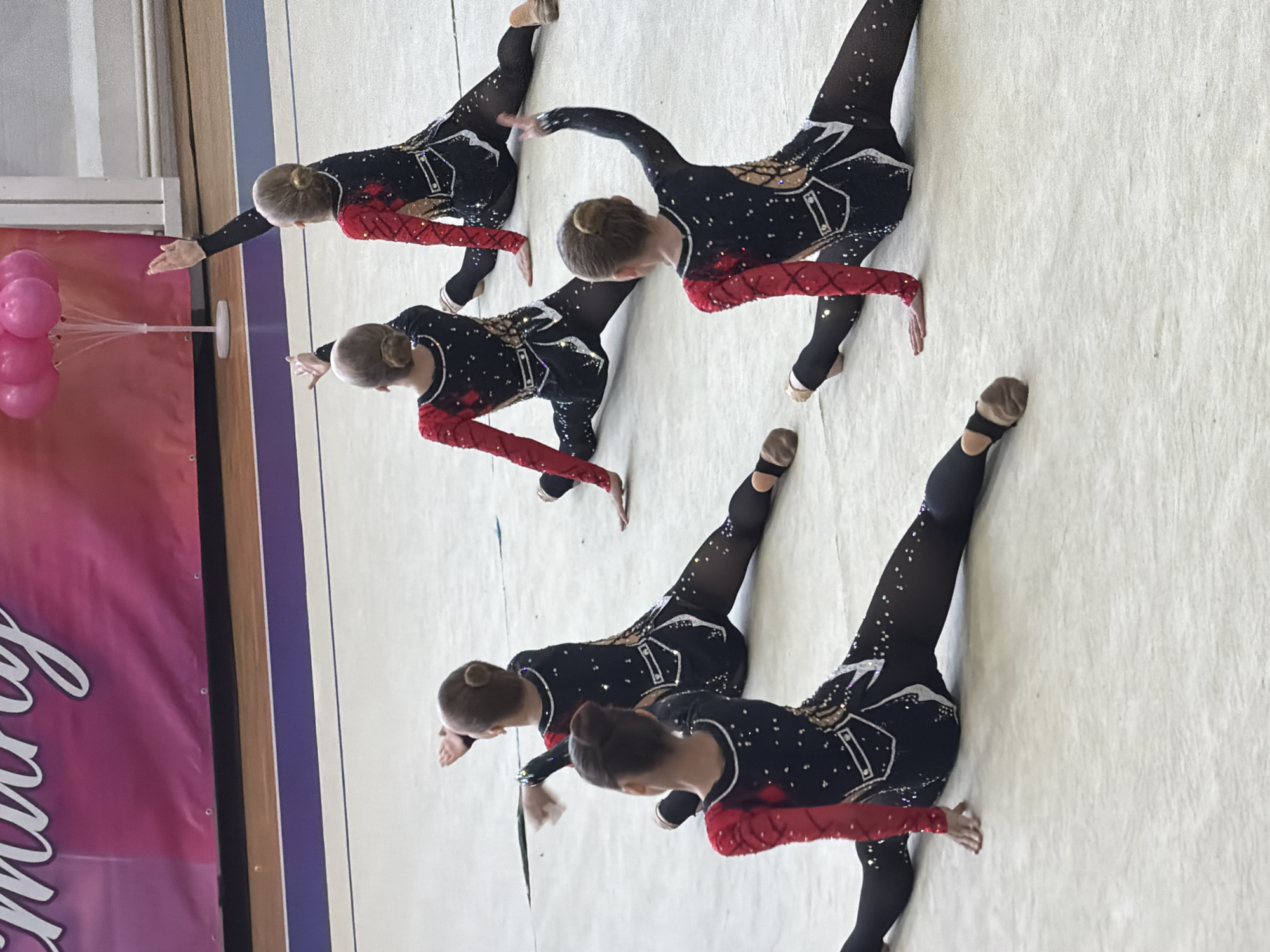 The image depicts five individuals performing a synchronized gymnastic routine on a light-colored mat. They are wearing black leotards with red sleeves and decorative patterns. Their hair is styled in buns. A pink banner is visible on the left side of the image, with unidentifiable white text. The location is unidentified.