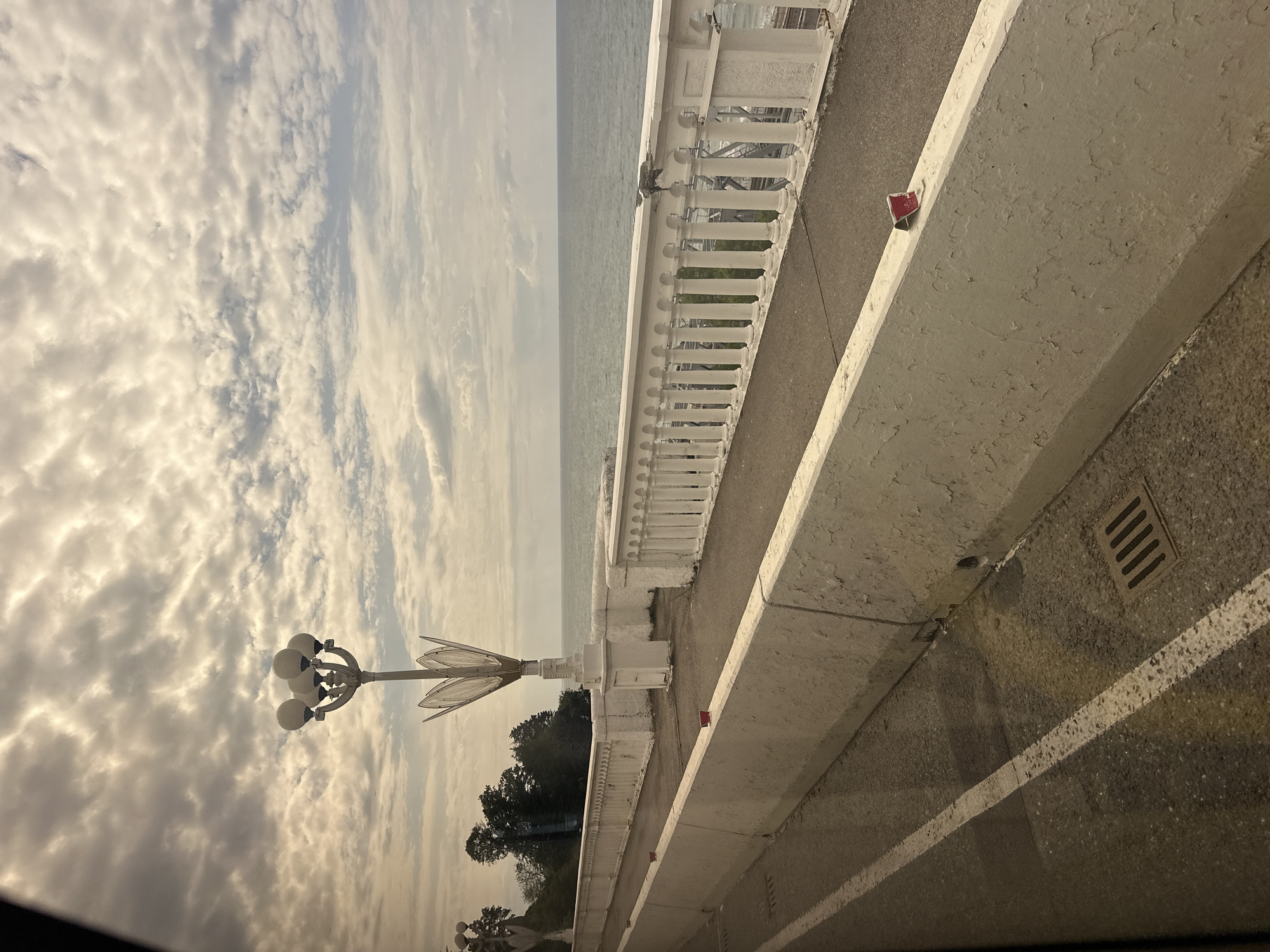 The image captures a scenic view of the coastline in Sochi, Russia, likely along the Arboretum or a similar promenade. The foreground features a grey asphalt road with a white lane marker and a concrete barrier. The barrier is painted white but shows signs of wear and tear, with peeling paint and what appear to be small red objects affixed to its top. A metal drain grate is visible on the pavement near the barrier.
Beyond the barrier, a paved walkway runs parallel to the sea. This walkway is bordered by a decorative white balustrade with ornate columns, characteristic of seaside promenades. Two tall, stylized streetlights, resembling stylized flowers with multiple globular lamps, stand along this walkway. The background is dominated by the vast expanse of the Black Sea, meeting a cloudy sky with patches of sunlight breaking through. Lush green trees can be seen on the left, suggesting proximity to a park or hillside. The overall scene conveys a somewhat overcast but picturesque coastal atmosphere.