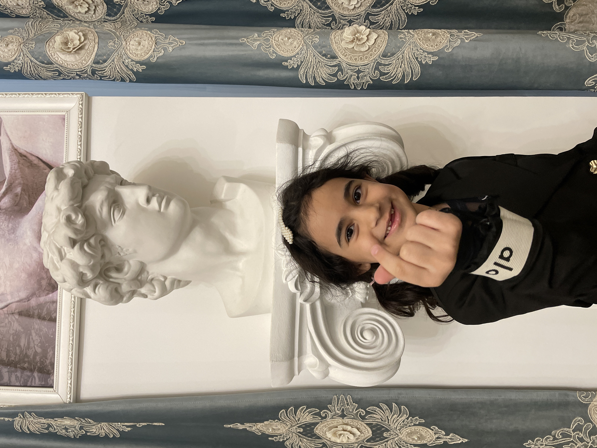 An upright indoor portrait features a young girl, a classical bust sculpture, and ornate decor. The central subject is a smiling young girl with dark hair, looking directly at the camera. She wears a black long-sleeved top and a pearl-studded hair clip. Her right hand is raised, making a finger heart gesture. On the black sleeve of her top, the white text "alo" is visible, oriented vertically.
To her left, a white bust sculpture, resembling a classical Greek or Roman figure like David, is prominently displayed on a white decorative architectural element that appears to be a corbel or console. Behind and slightly to the left of the bust, a framed artwork with soft purple and pink tones is partially visible. The background wall is white.
The upper and lower parts of the frame are adorned with luxurious, teal-blue fabric drapery featuring elaborate gold or beige embroidered patterns with floral and curvilinear motifs, some including raised or beaded details. The overall setting suggests an elegantly decorated indoor space, possibly a home, with a blend of classical art and rich textiles. The lighting is bright and even, indicating an indoor environment, likely during the day. The girl appears happy and engaged, adding a cheerful element to the refined setting.