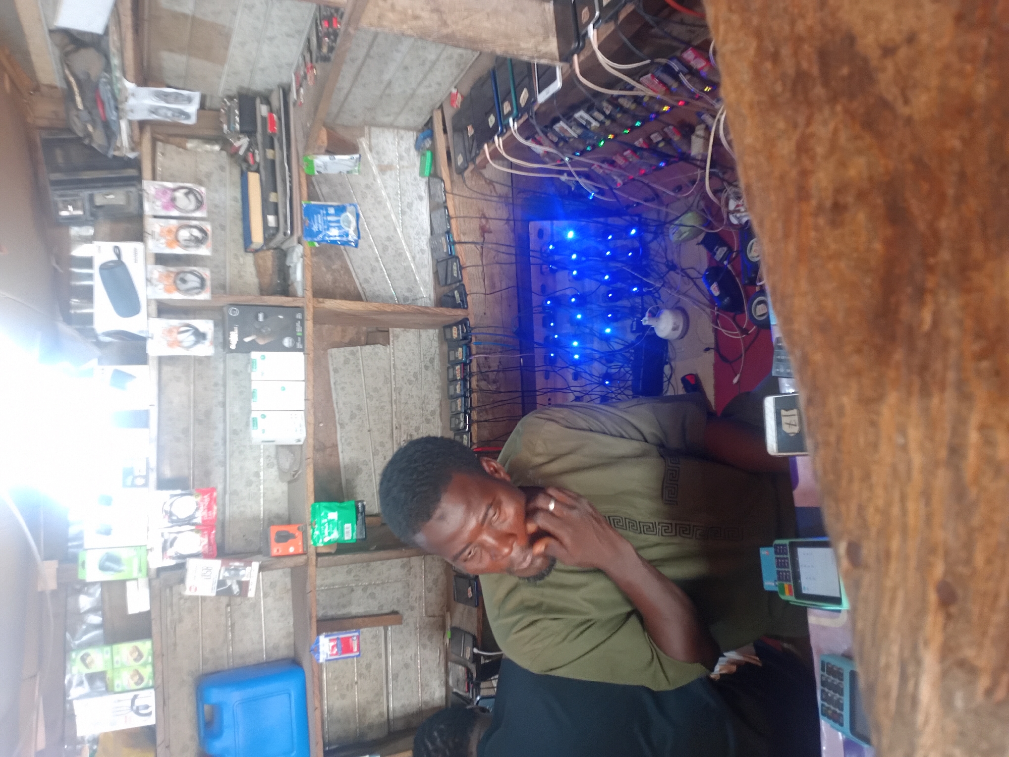 A man in an olive green patterned shirt sits behind a counter in a small shop. He is touching his face with his right hand. The shop has shelves stocked with electronic accessories, including headphones, speakers, and chargers. Multiple charging devices are plugged into a central board with blue lights. A second person can be seen in the lower part of the image. The location is Unknown, Unknown.