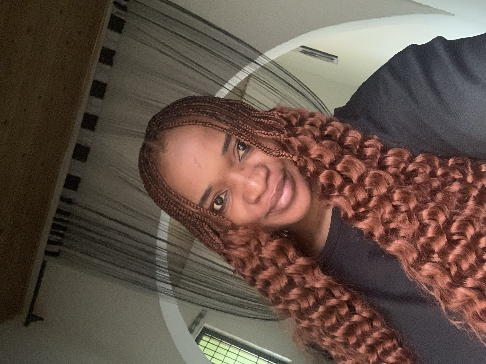 A close-up selfie of a woman with auburn box braids wearing a dark-colored shirt. The background features a window with a view of greenery and a white architectural arch with a hanging black curtain-like decoration. The photograph does not provide verifiable location information related to Barakin Akawo, Nigeria.